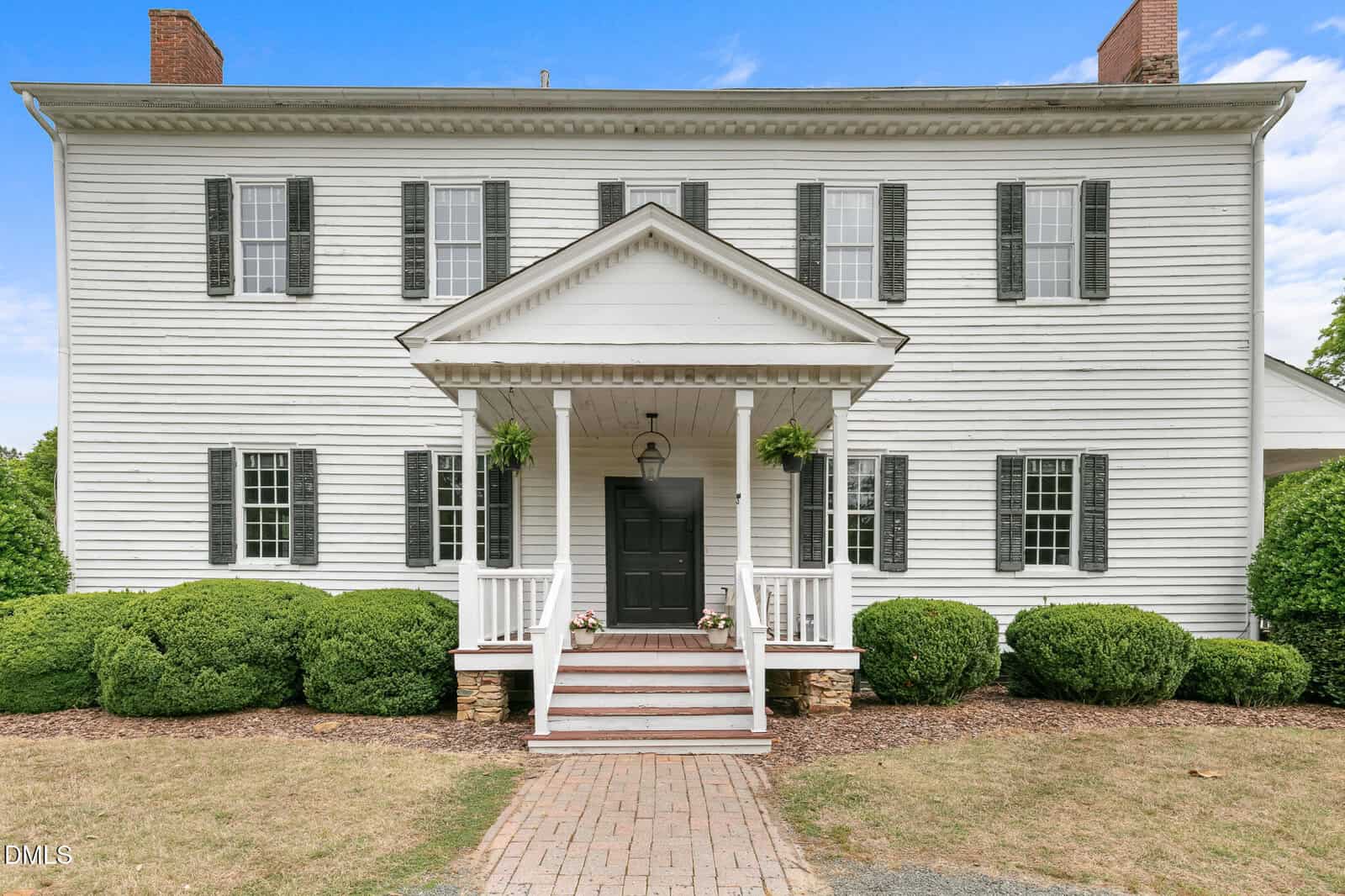 A two-story white colonial-style house with black shutters, a central porch with columns, potted plants, and manicured bushes along the front, under a blue sky.