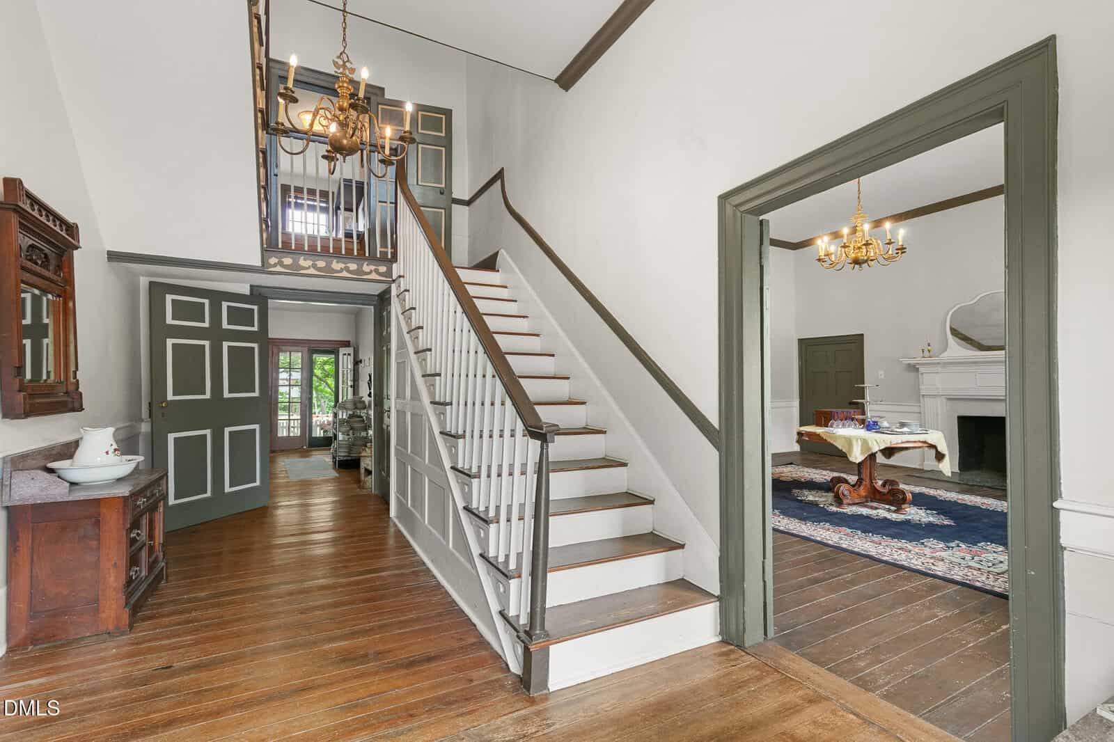 Bright entryway of a historic home with wooden floors, a white staircase with dark trim, a chandelier, and an open doorway leading to a room with a fireplace, chandelier, and area rug.
