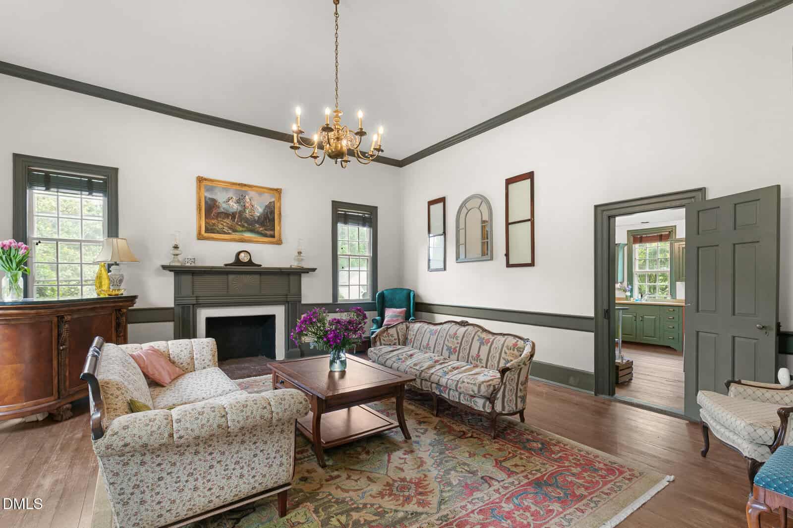 Bright, elegant living room with vintage sofas, a floral armchair, coffee table, fireplace, chandelier, and wall art. Large windows let in natural light; an open door reveals a green kitchen beyond.
