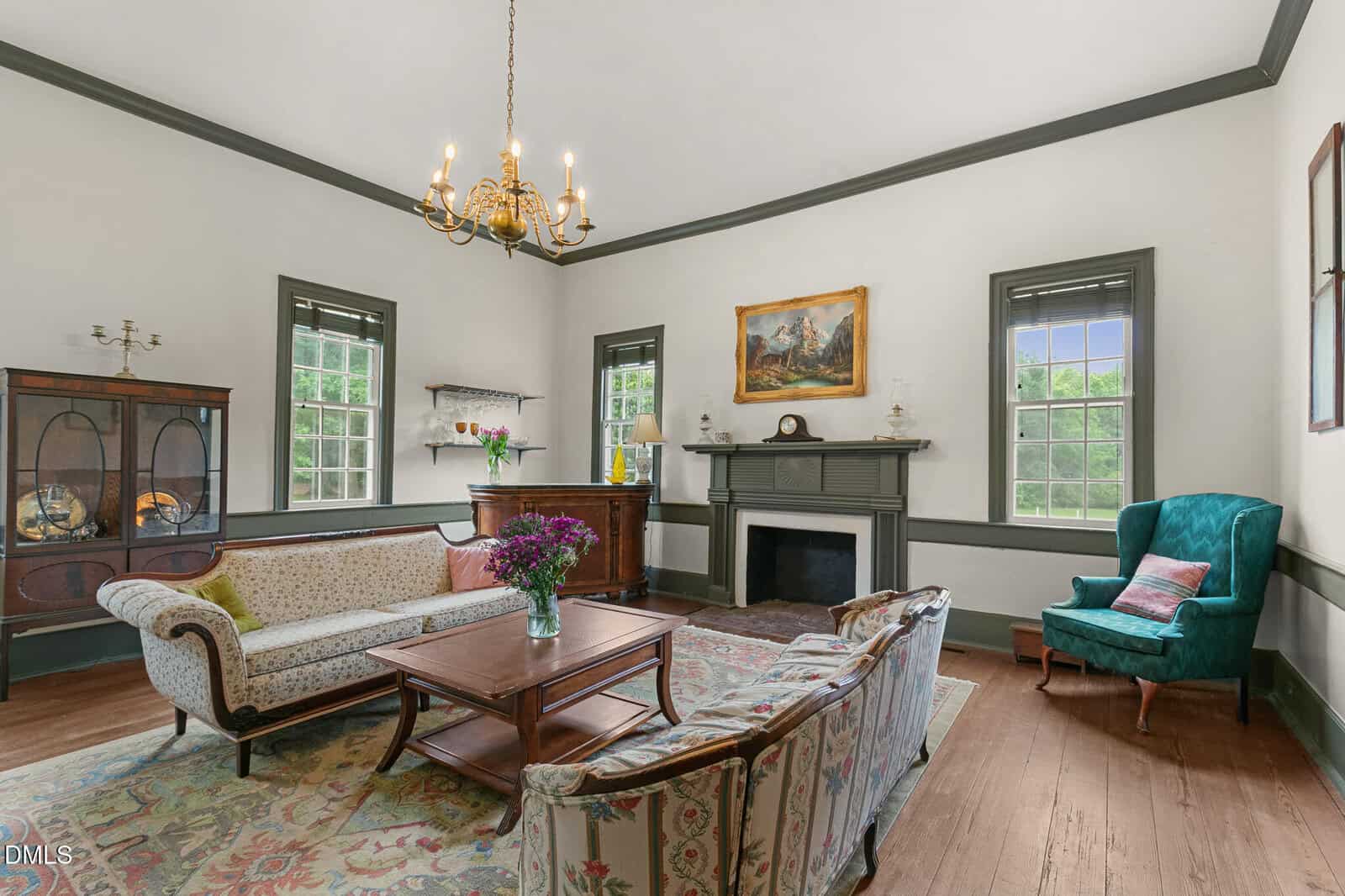 Bright living room with vintage sofas, wooden coffee table, fireplace, and chandelier. Green wingback chair and floral rug add charm, with artwork and large windows completing the classic decor.
