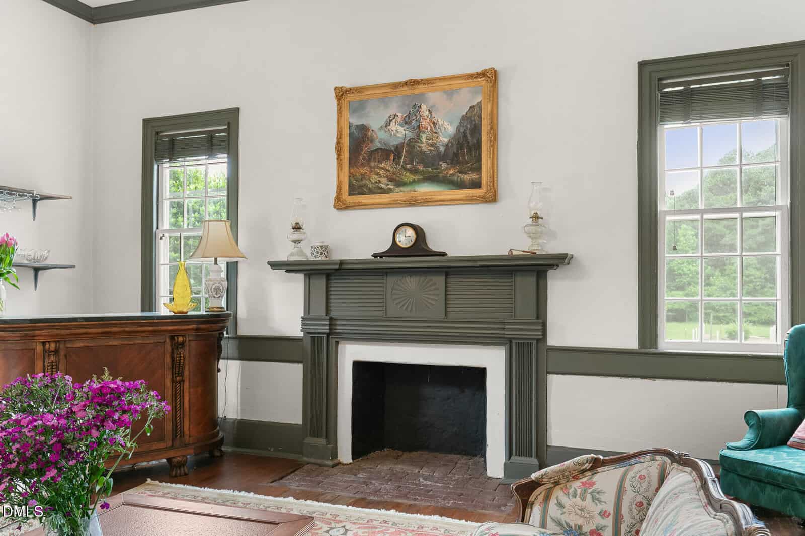 A classic living room with a gray-trimmed fireplace, a vintage mantel clock, oil lamps, landscape painting, large windows, wooden sideboard, sofa, and a floral bouquet on a table.
