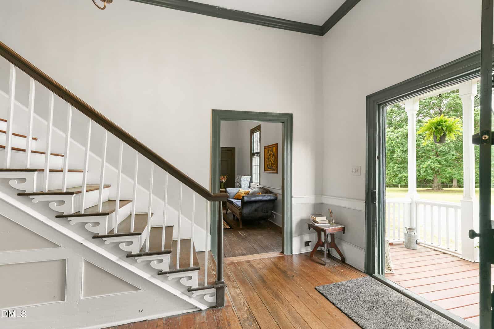 Bright entryway with wooden stairs, open door leading to a porch, and a view into a living room with a dark couch. Hardwood floors and white walls with gray trim throughout.
