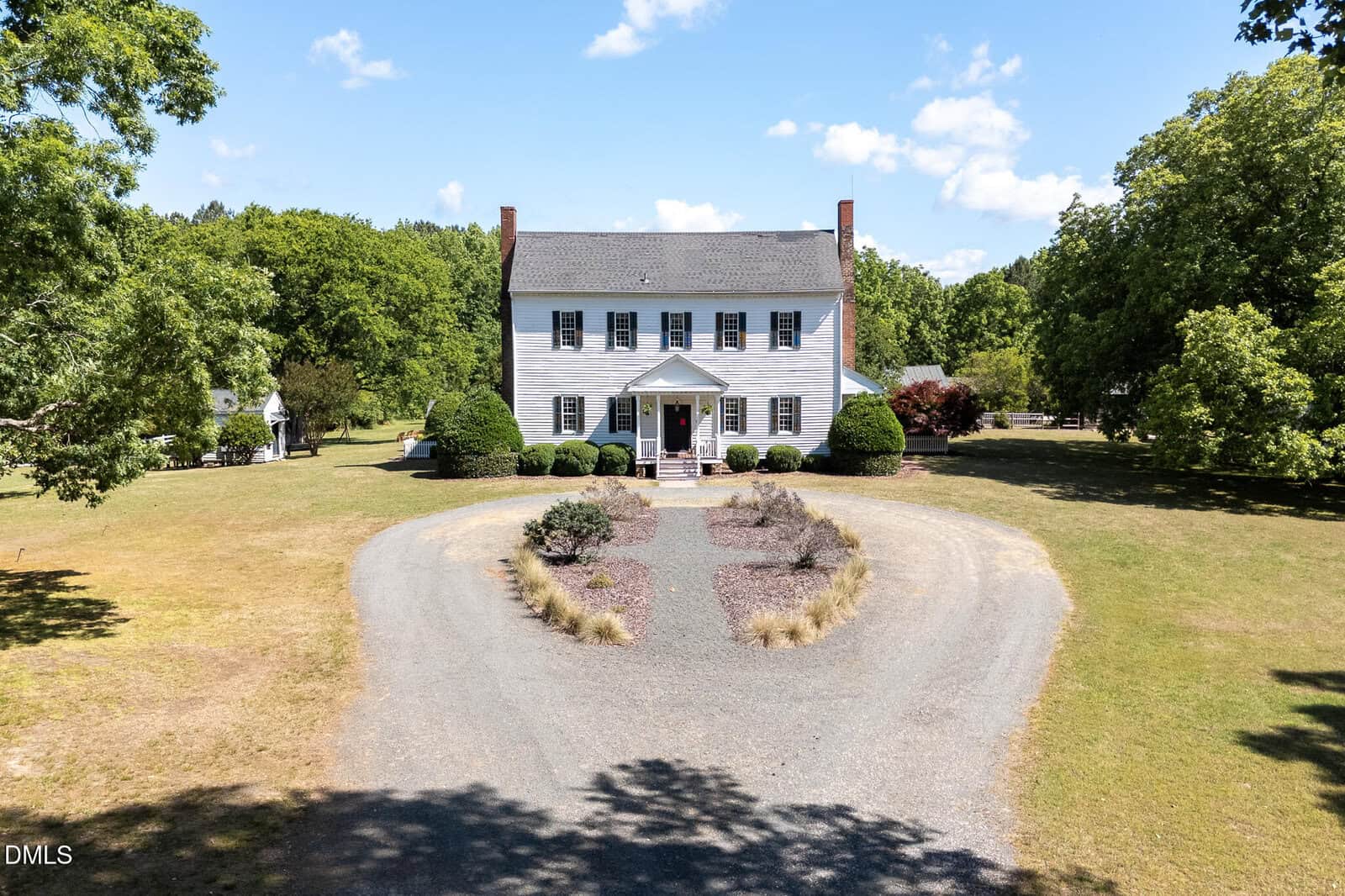 A large, two-story white colonial-style house with a gray roof sits at the center of a circular gravel driveway, surrounded by lawn, trees, and landscaped bushes under a blue sky with scattered clouds.