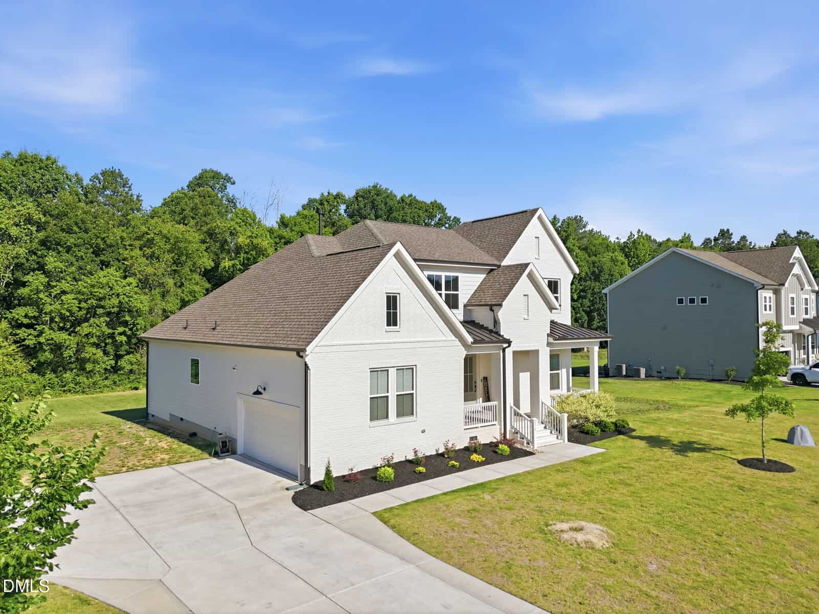 A modern two-story white house with a covered front porch, attached garage, and neatly landscaped yard. The home sits on a spacious lot with green grass and is bordered by trees and neighboring houses under a clear blue sky.