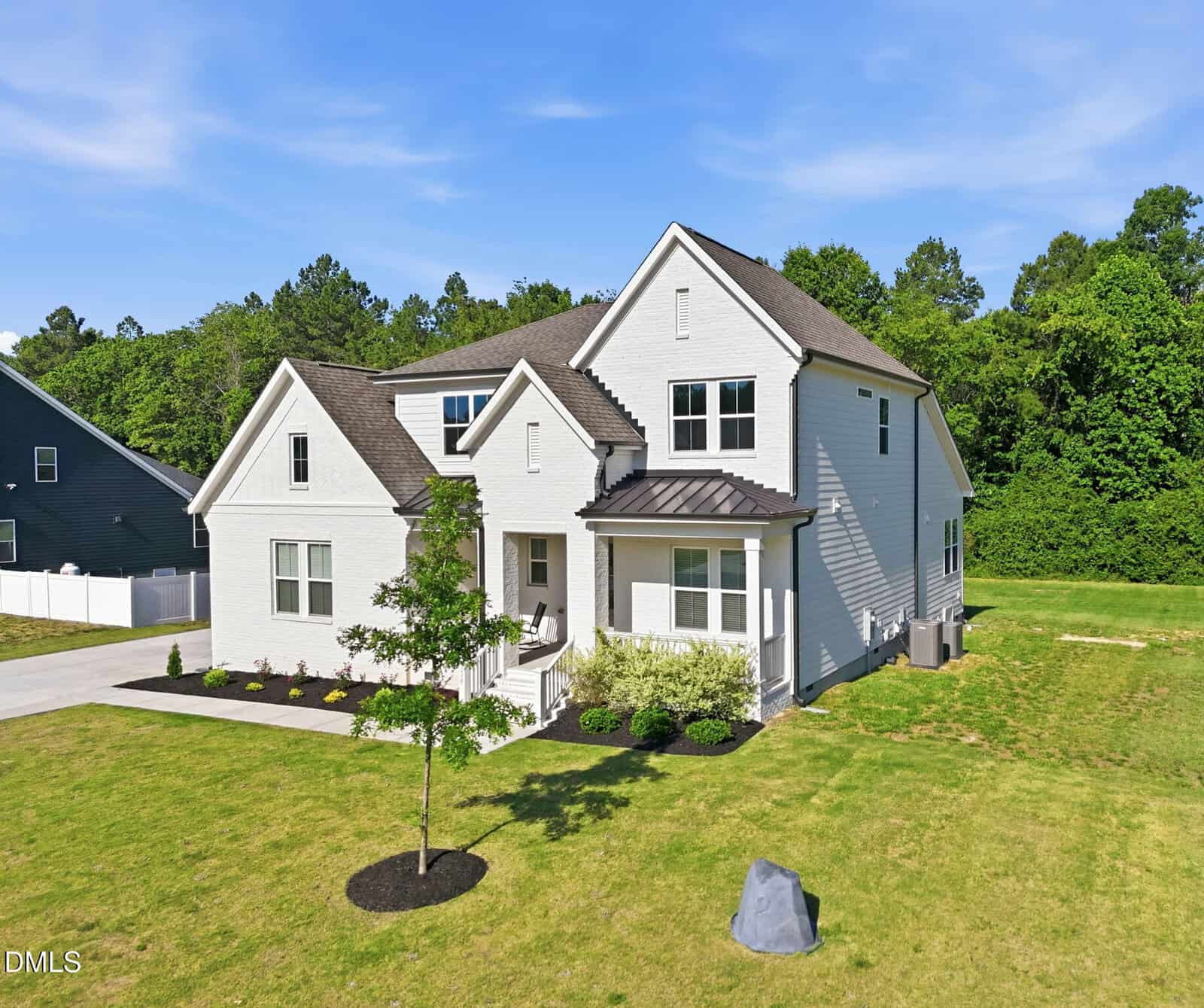 A modern two-story white house with dark roof shingles, a small front porch, neatly landscaped yard, and a tree in the front, set against a backdrop of trees and blue sky on a sunny day.
