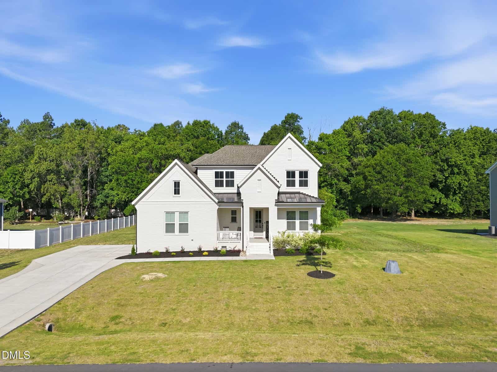 A large, modern two-story white house with a front porch, driveway, and well-maintained lawn, surrounded by trees under a blue sky.