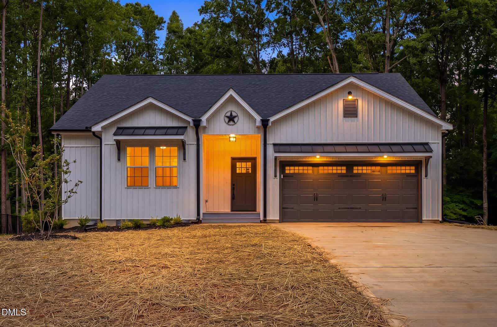 A modern single-story house at 103 Yuma Drive in Louisburg features white vertical siding, dark trim, and a double garage. Warm lights illuminate the porch and garage, with trees and a freshly mulched yard surrounding the home.
