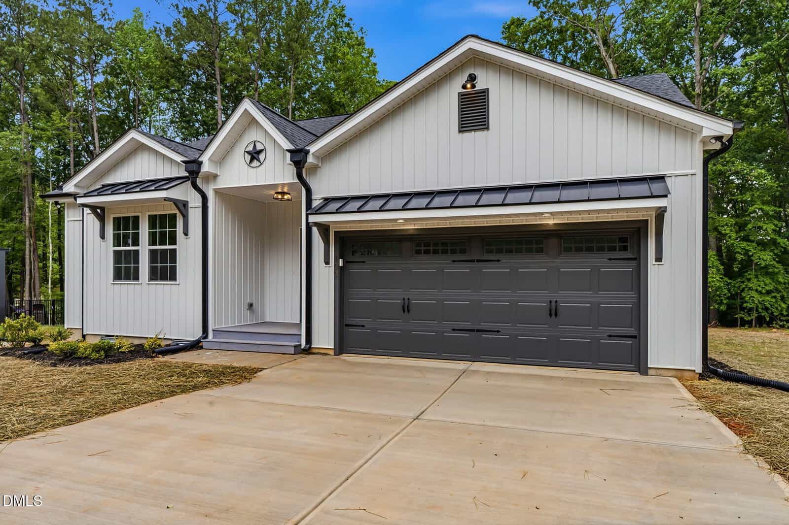 Modern single-story house with white vertical siding, black trim, and a large dark gray two-car garage. The home has a small front porch, concrete driveway, and is surrounded by trees and fresh landscaping.