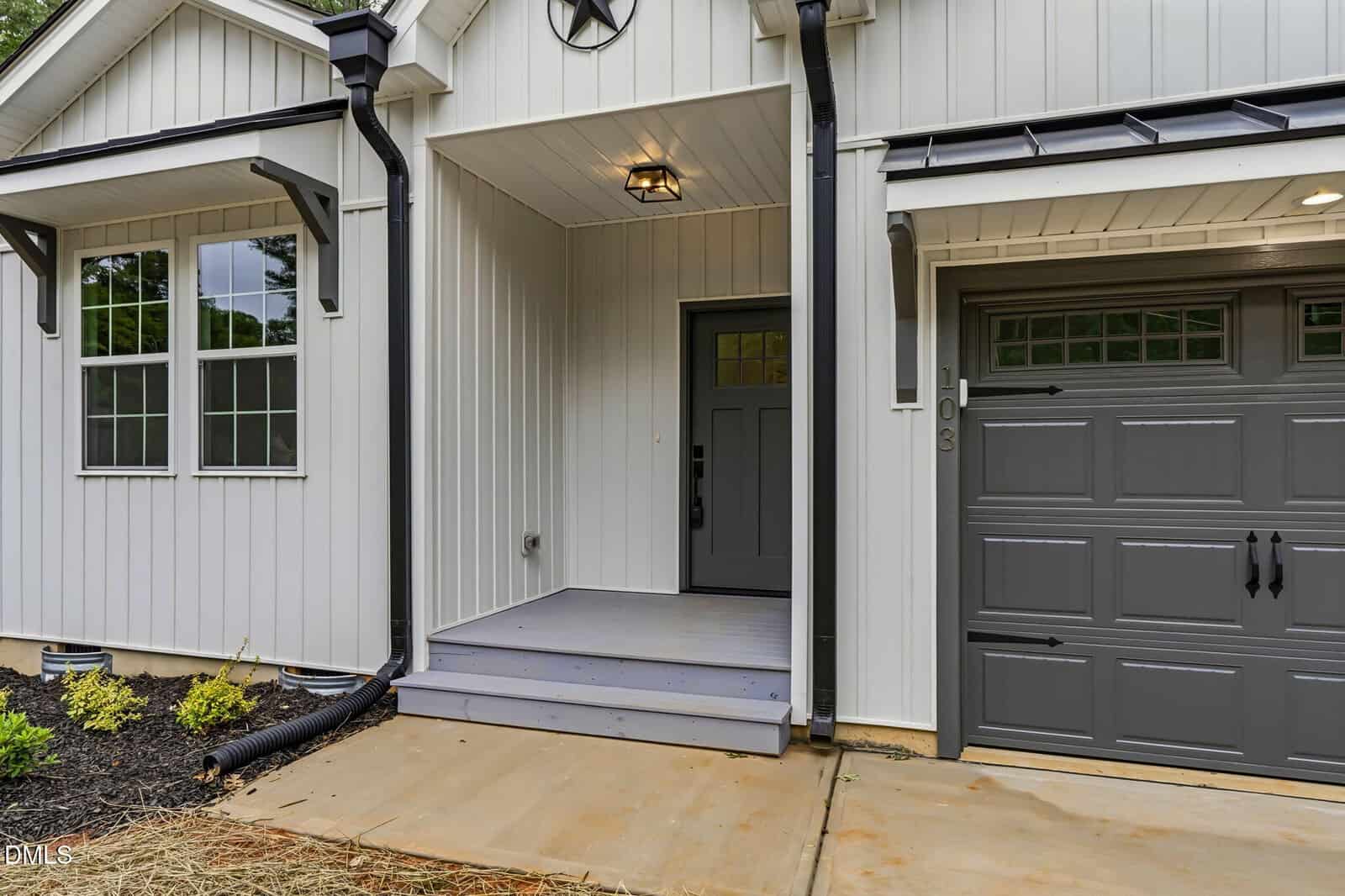 A modern house exterior features white vertical siding, a gray front door with a small porch, black trim, and a gray garage door. There are two windows to the left and minimal landscaping with mulch and small plants.
