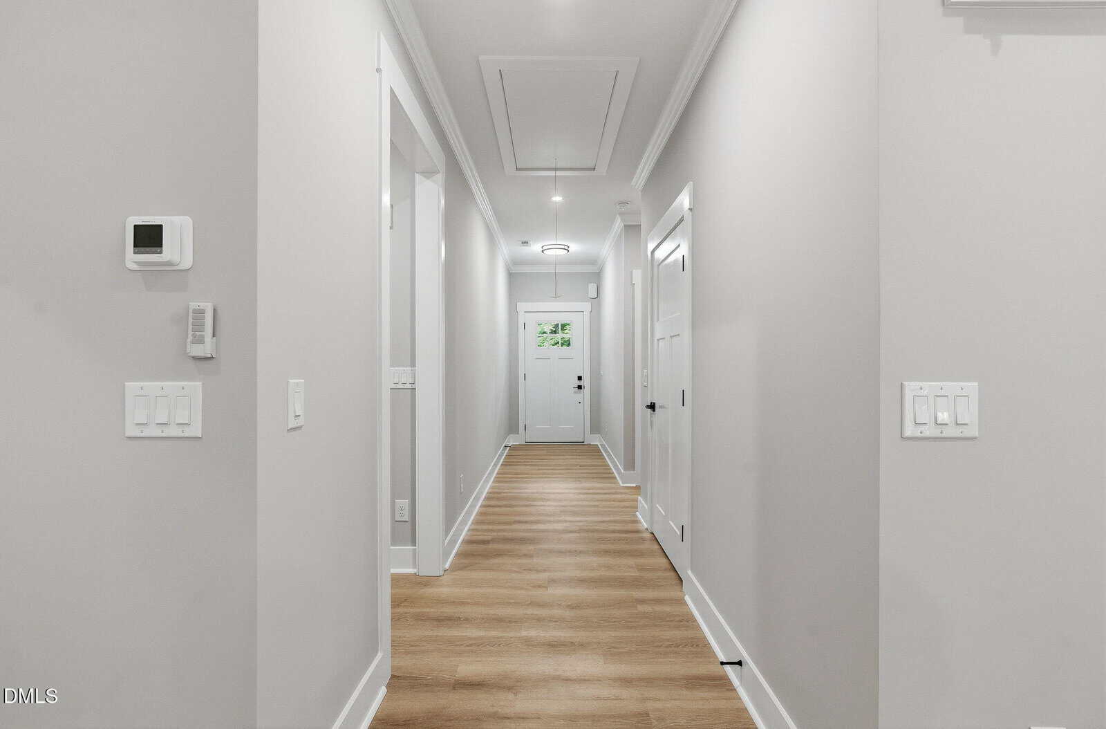 Long, bright hallway with light-colored walls, high ceiling, wood floor, white trim, multiple doors, wall switches, and a thermostat. A glass-paneled door and a window are visible at the far end.