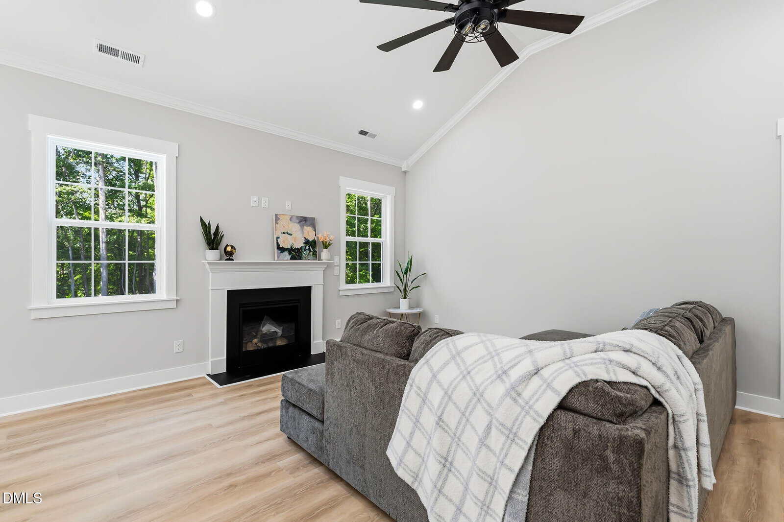 A bright living room with light wood floors, gray walls, a gray sectional sofa with a plaid throw, a white mantel fireplace, green plants, and large windows letting in natural light.