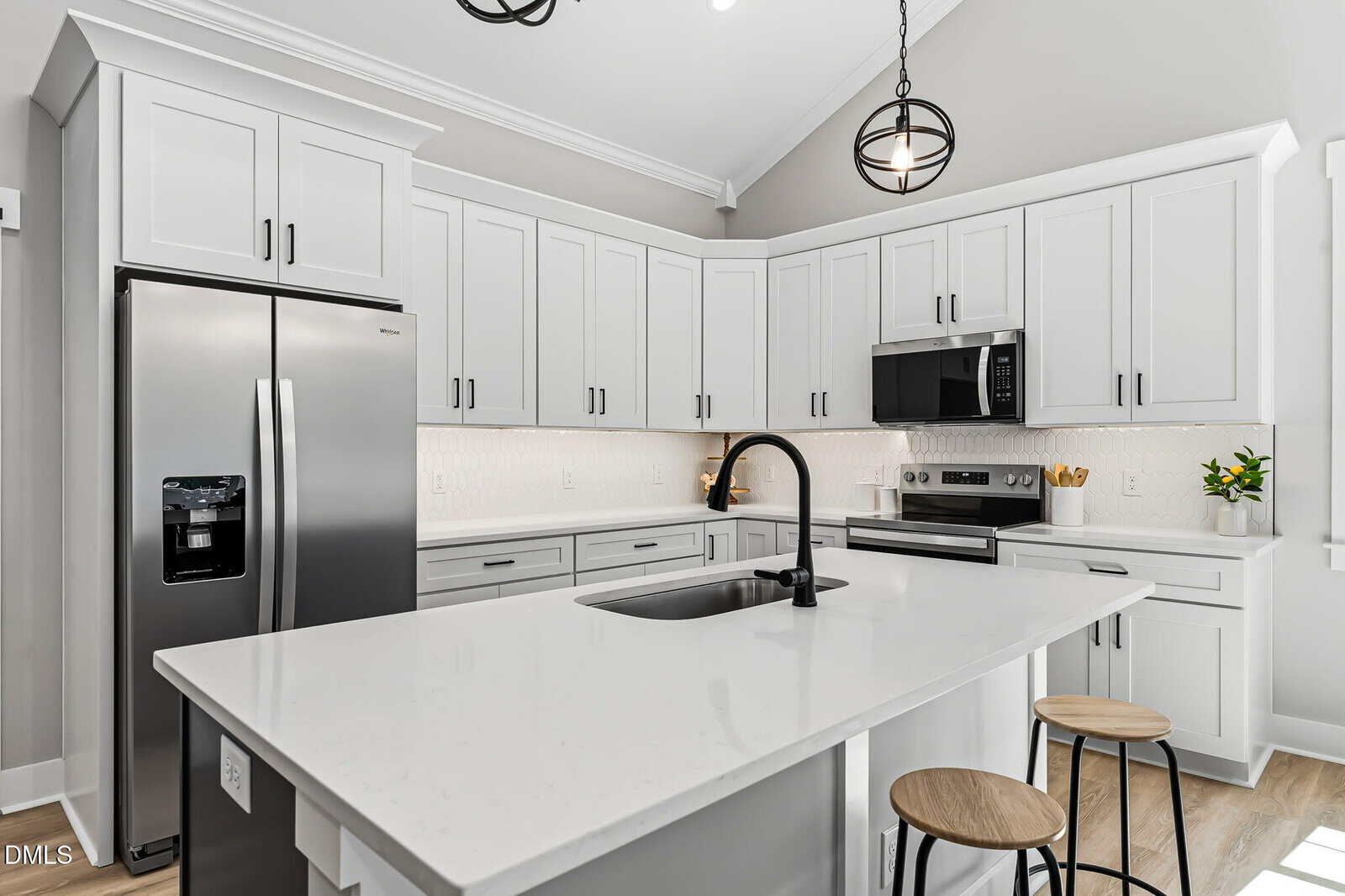 Modern kitchen with white cabinets, stainless steel appliances, black fixtures, and a white island with a sink and two wooden stools. Geometric pendant lights hang from the vaulted ceiling.