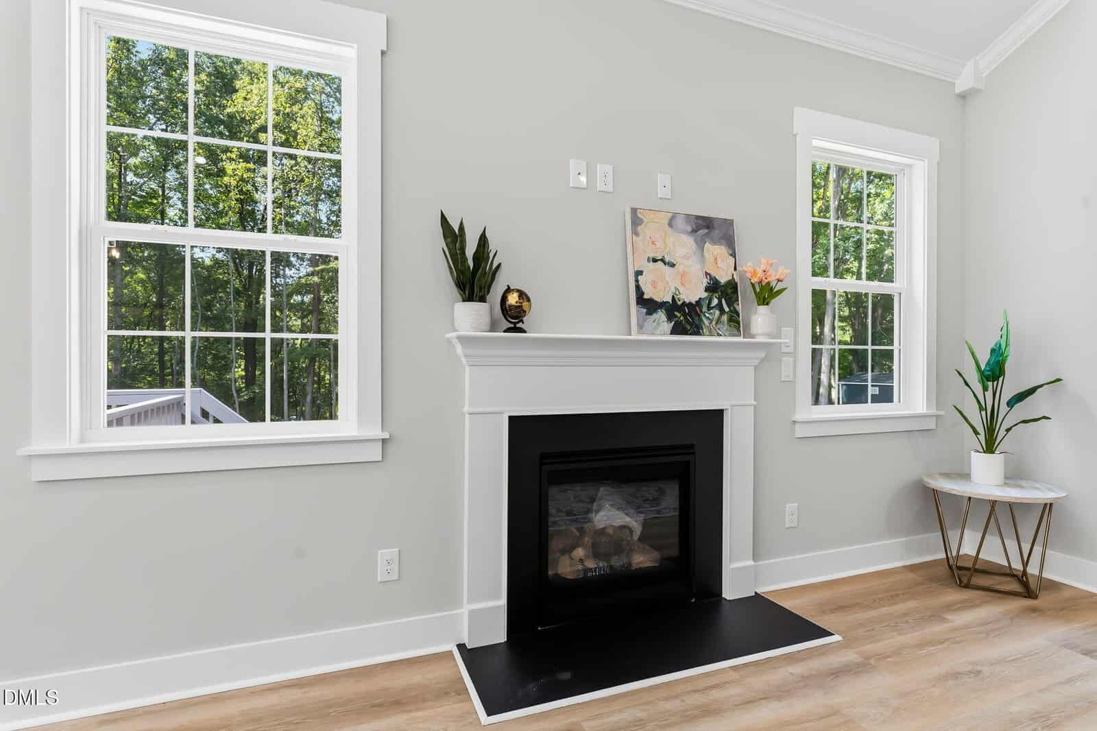 Bright living room with a white fireplace, plants, and flower art above the mantel. Two large windows let in natural light. Light wood floors and a small round side table on the right complete the space.