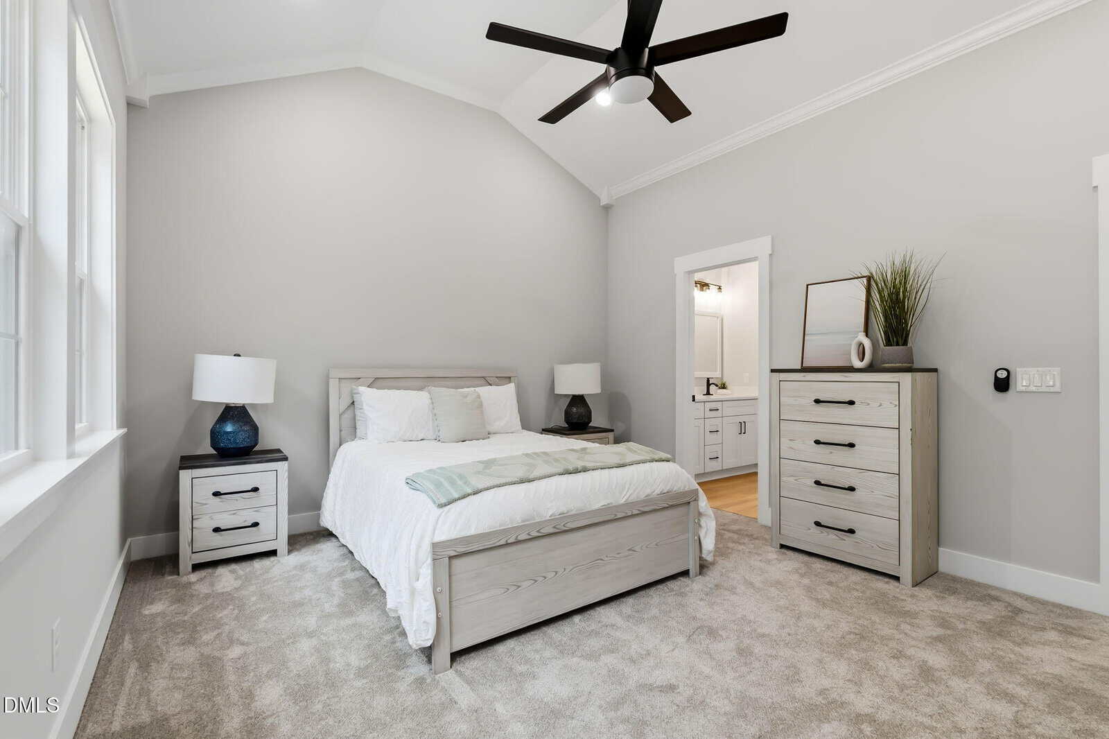 A modern bedroom with a bed, two nightstands, a dresser, neutral walls, carpeted floor, ceiling fan, lamps, and a doorway leading to a bathroom. Simple decor with plants and vases.