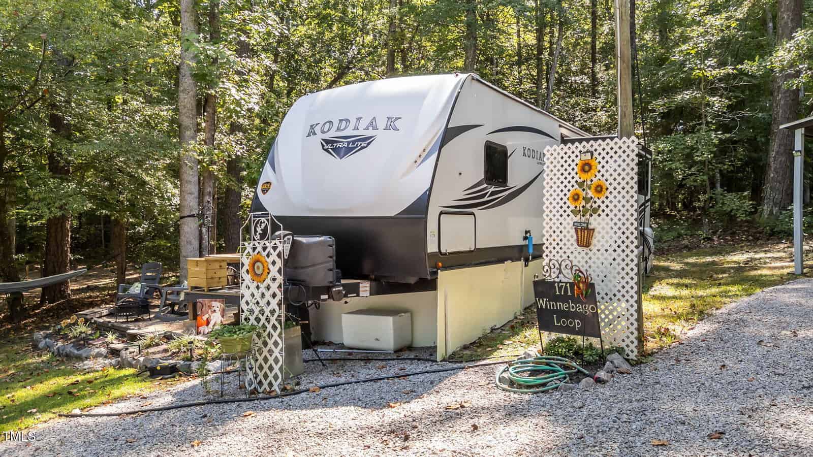 A white Kodiak travel trailer is parked on gravel in a wooded area, with sunflower-themed decorations and a sign reading "Winnebago Loop" near the front. Trees and greenery surround the campsite.