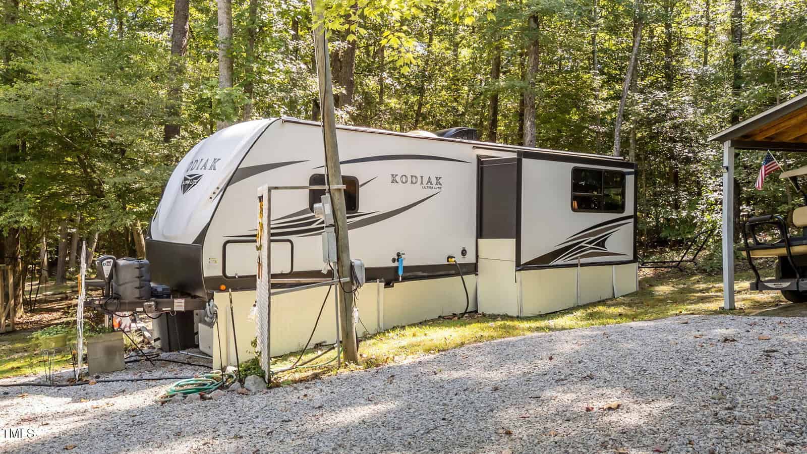 A white Kodiak travel trailer is parked on a gravel surface in a wooded area, with trees in the background and a small structure partially visible to the right.