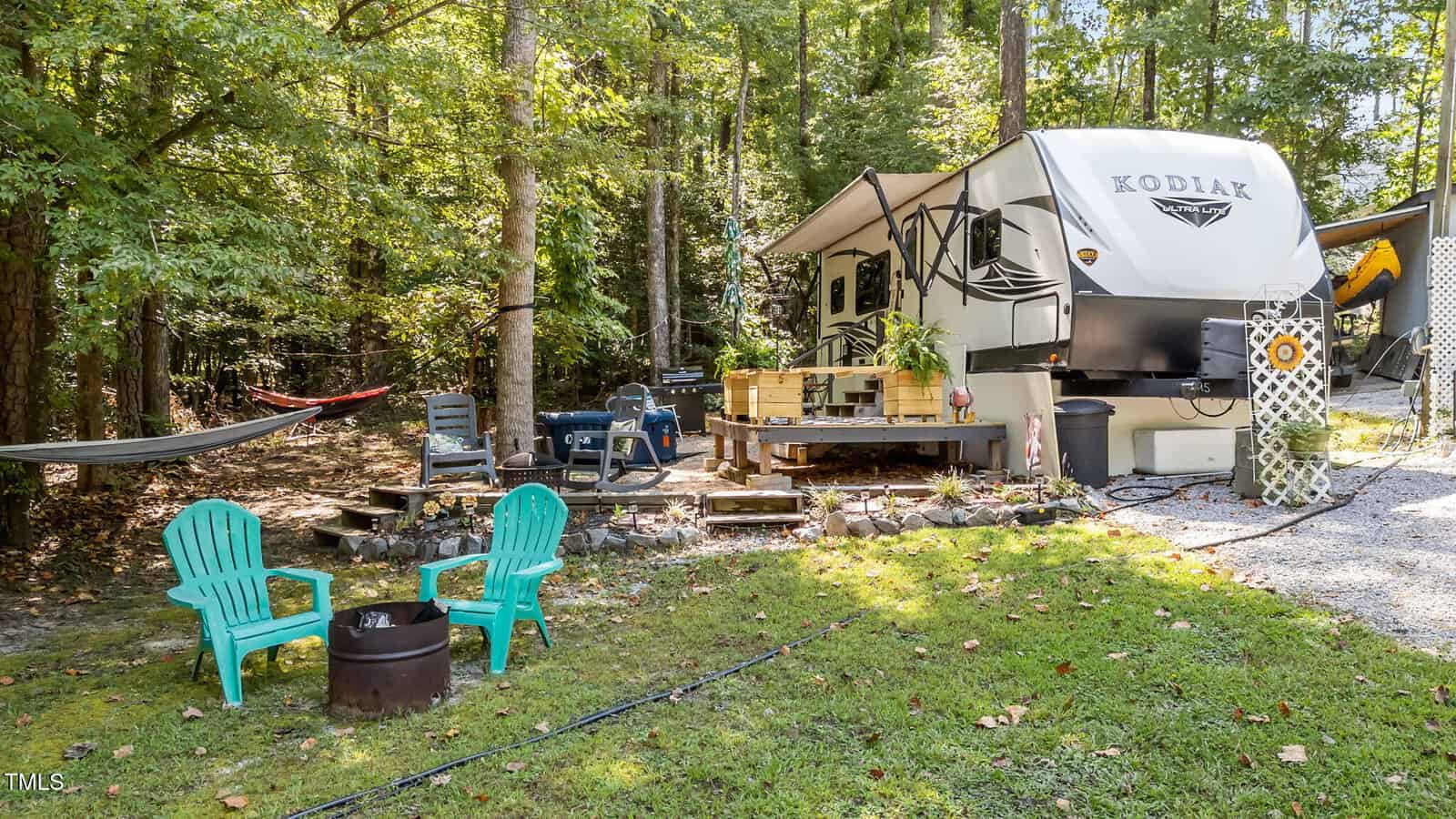 A large RV parked in a wooded area with a deck, outdoor seating, a hammock, two teal chairs around a fire pit, and various camping accessories. Trees surround the campsite, and grass covers the ground.