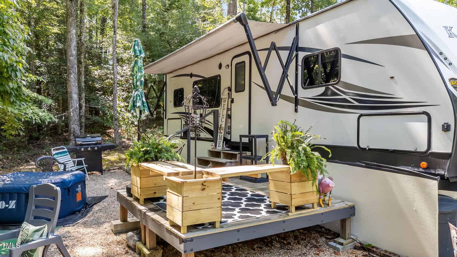 A cozy campsite with a beige RV, a small wooden deck with potted plants, a picnic table, outdoor seating, and a grill, surrounded by tall green trees and forest.