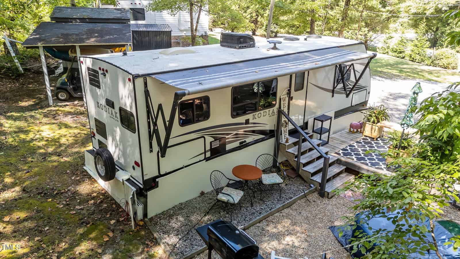 A large RV with outdoor seating, table, and stairs leading to a rug-covered patio is parked among trees on a gravel lot, with shade structures and greenery nearby.