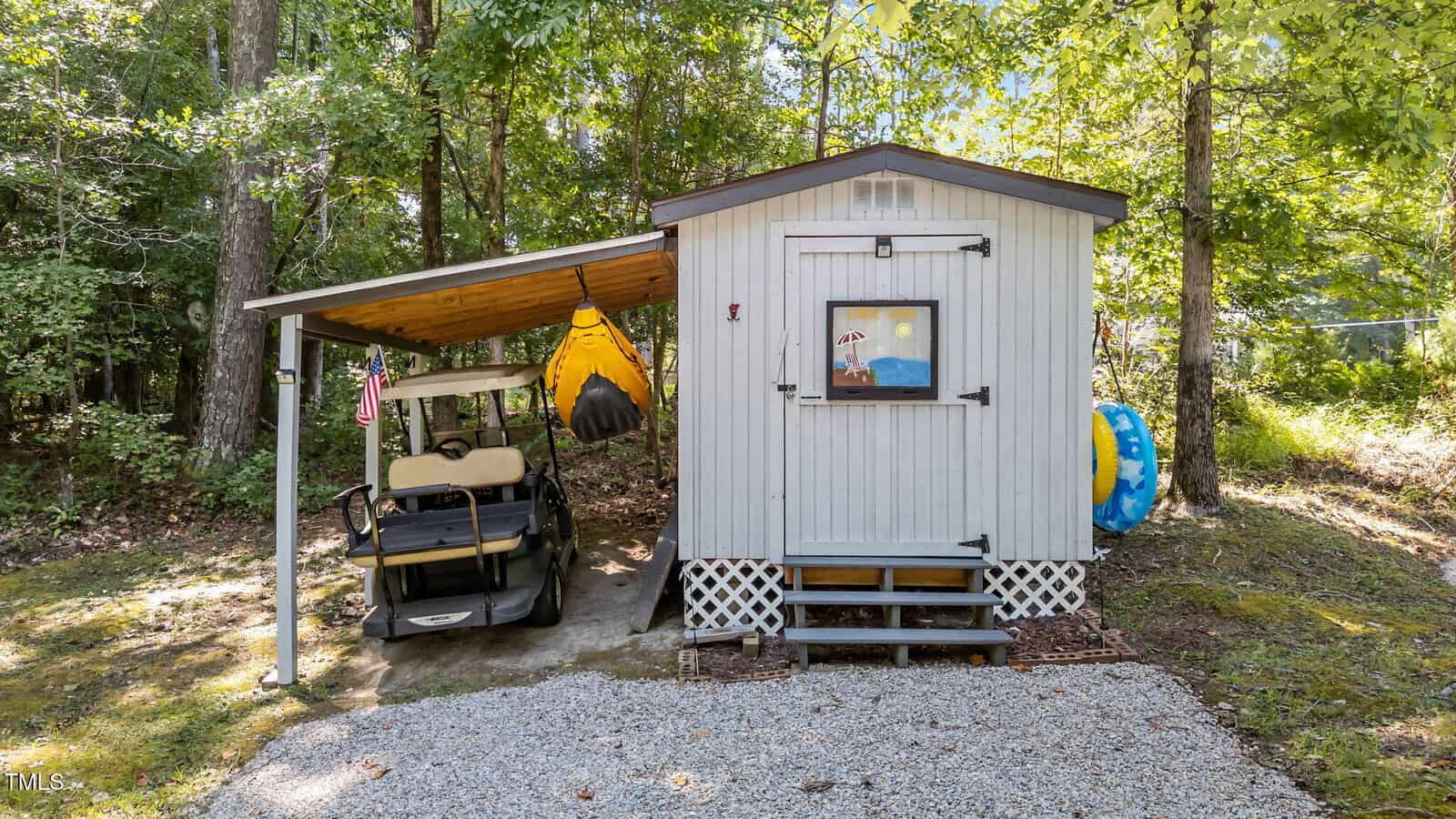 A small gray shed with a window stands in a wooded yard. A golf cart and two yellow kayaks are stored under the attached side roof. A round blue pool float leans against the shed. Gravel covers the ground in front.
