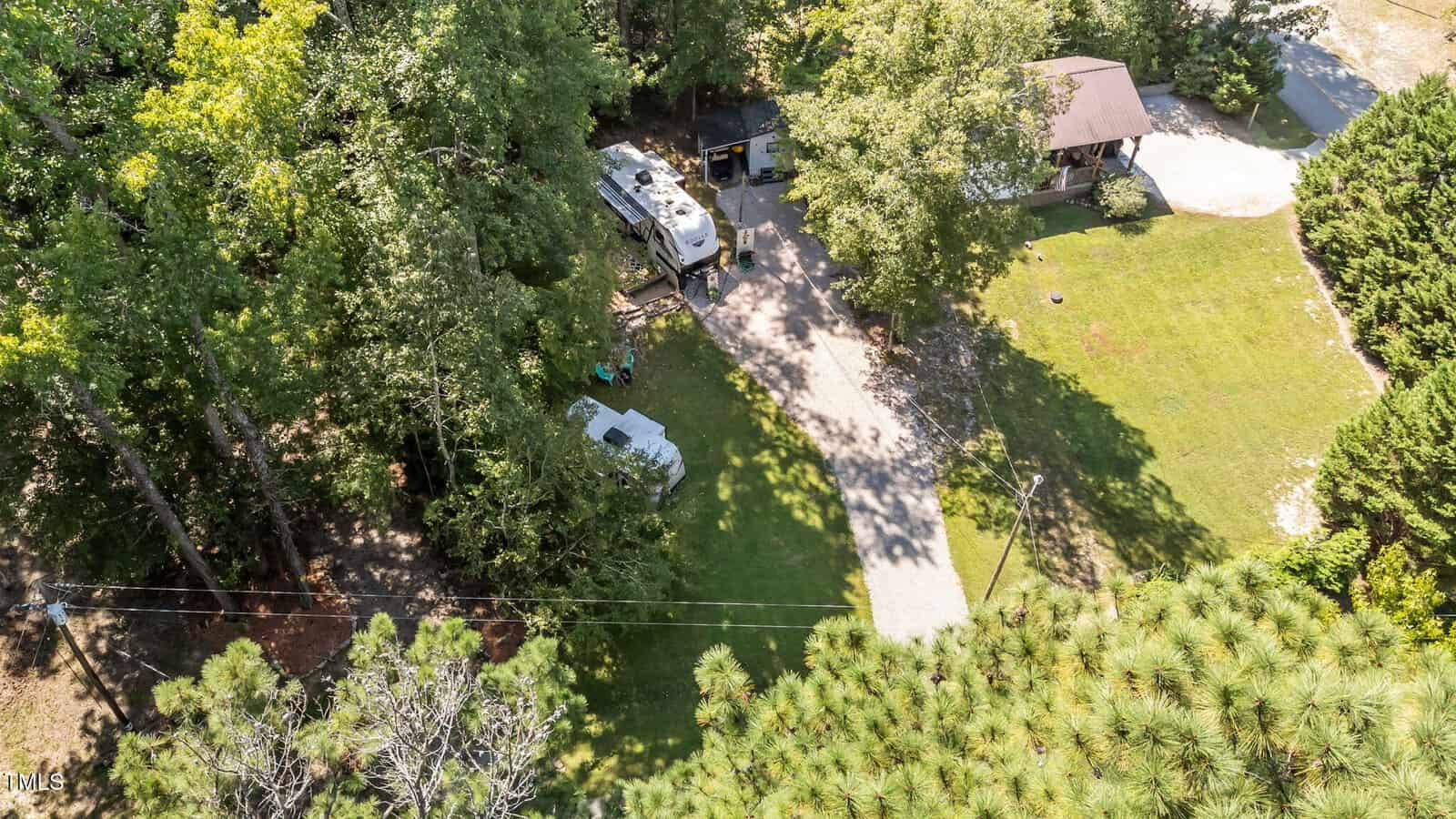 Aerial view of a house with a driveway, green lawn, trees, and two RVs parked near a wooded area. Sunlight creates shadows on the ground.