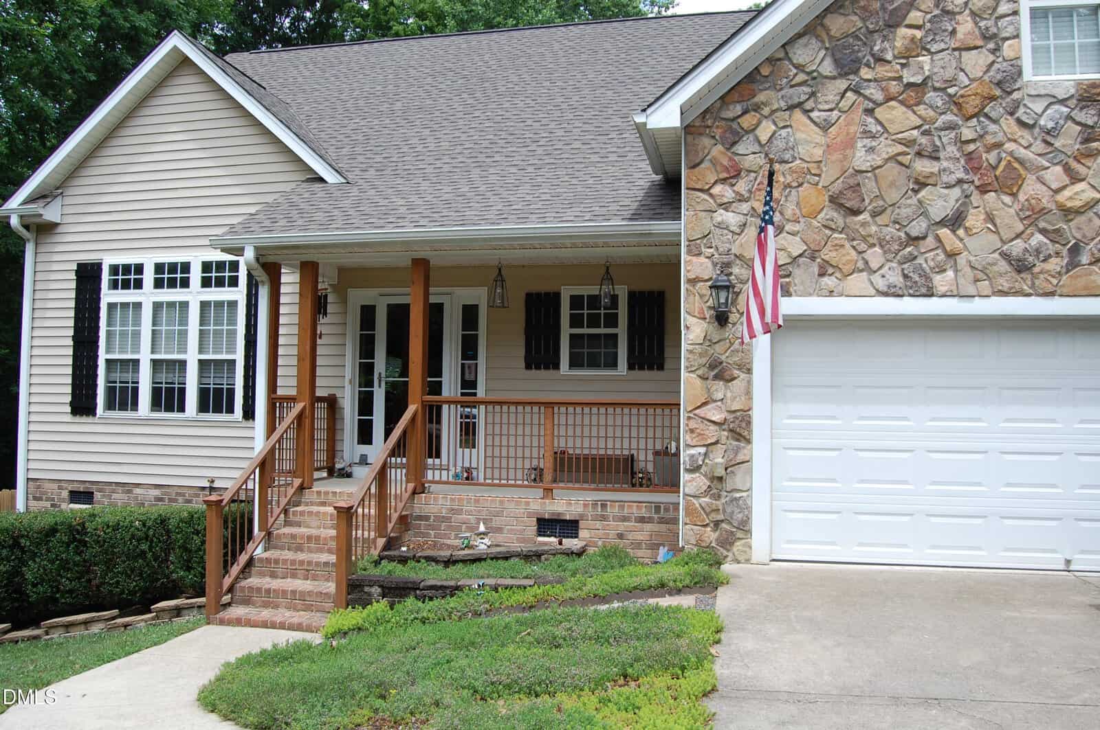 A tan house with a stone facade, white garage door, front porch with wooden railings, brick steps, black shutters, hanging lanterns, and a U.S. flag by the driveway, surrounded by green shrubs.