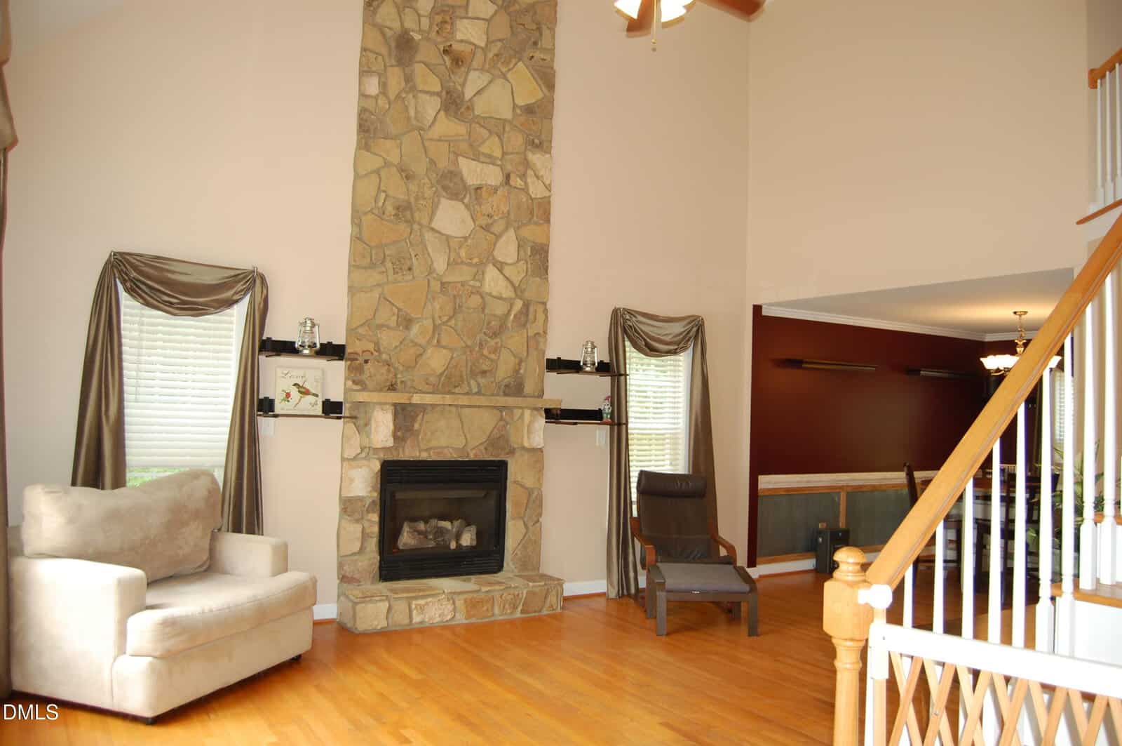 A living room with hardwood floors, a tall stone fireplace, two windows with drapes, a beige armchair, a dark accent chair, and a view of a dining area with dark red walls and built-in shelves.