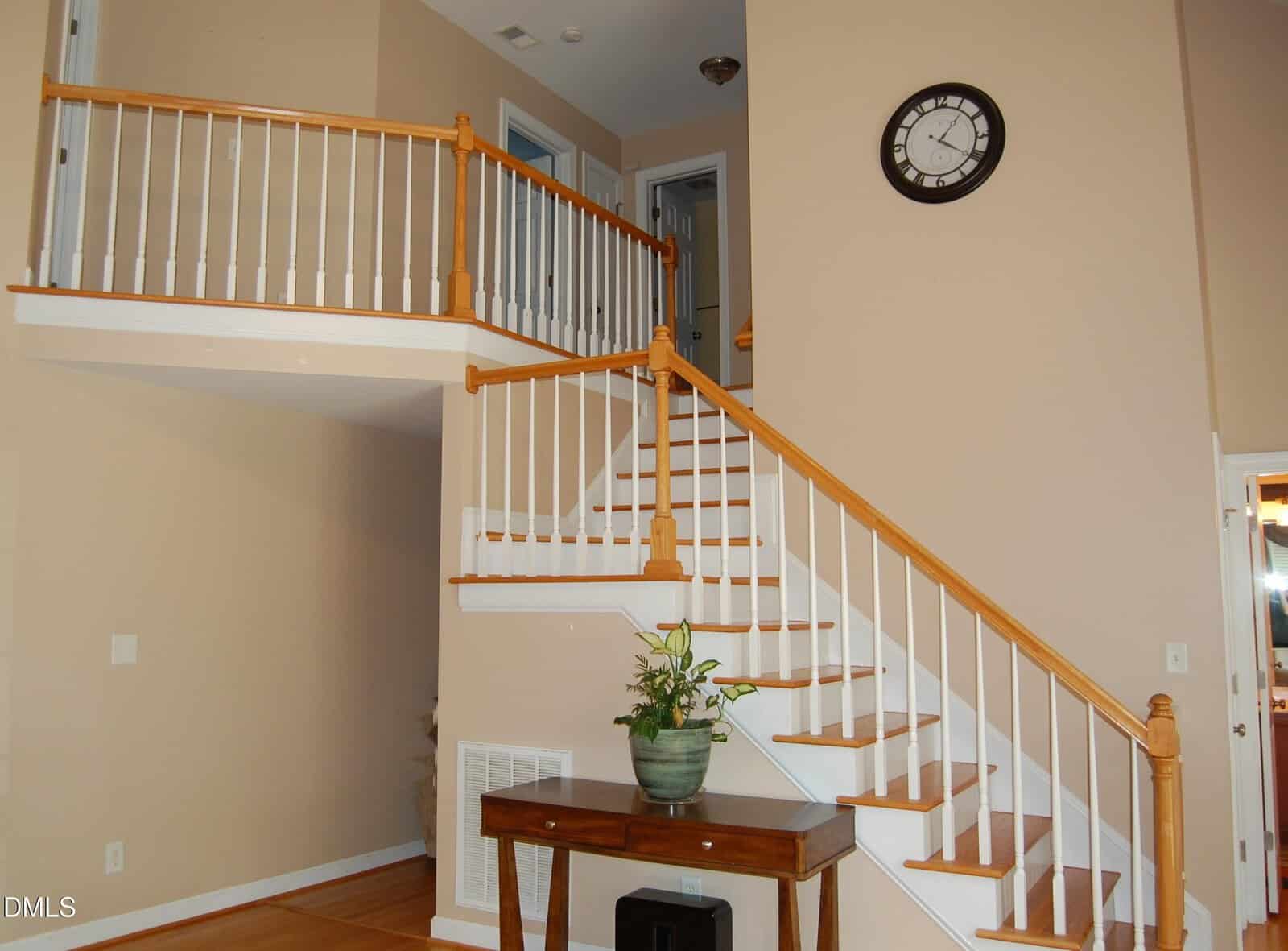 A staircase with wooden steps and white balusters leads to an upstairs hallway. A potted plant sits on a table below the stairs. A round wall clock is mounted above the landing. Walls are painted beige.
