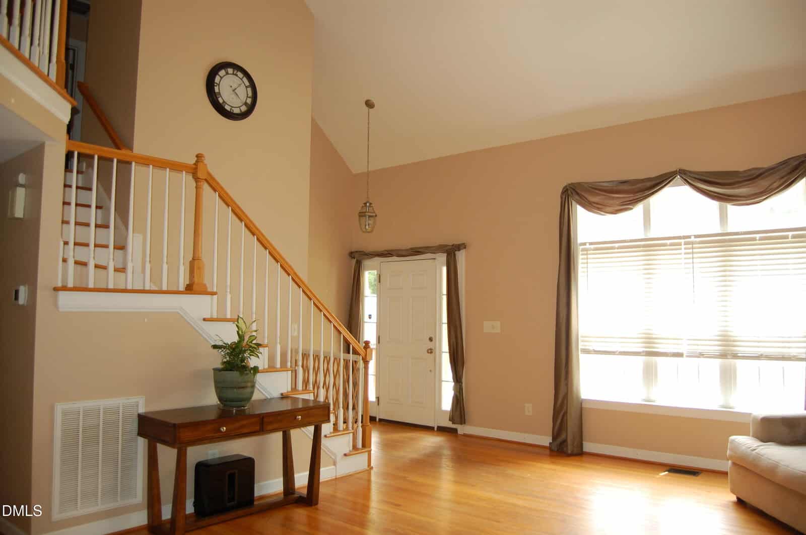 Bright foyer with hardwood floors, a large window with drapes, white front door, staircase with wooden railing, side table with a plant, and a wall clock above. Natural light fills the space.