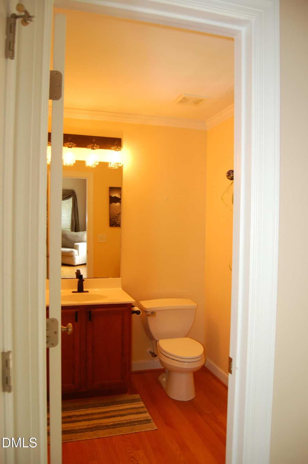A small bathroom with a wooden vanity, sink, toilet, wall mirror, light fixture above, and a striped rug on the hardwood floor. A painting is on the wall, and a doorway leads to another room.