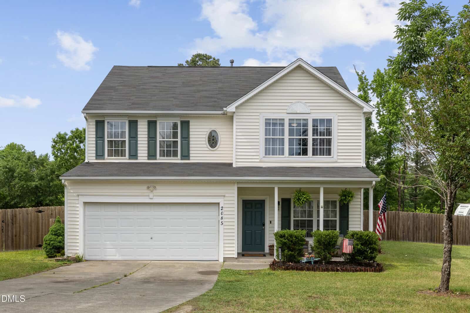 Two-story suburban house with light siding, dark shutters, and a gray roof. Features a two-car garage, green front door, porch with plants, small lawn, and an American flag. Trees and a wooden fence surround the yard.