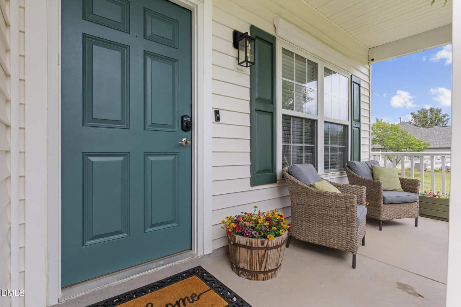A front porch with a teal door, two wicker chairs with cushions, a barrel planter with colorful flowers, a welcome mat, and large windows on a cream-colored house.