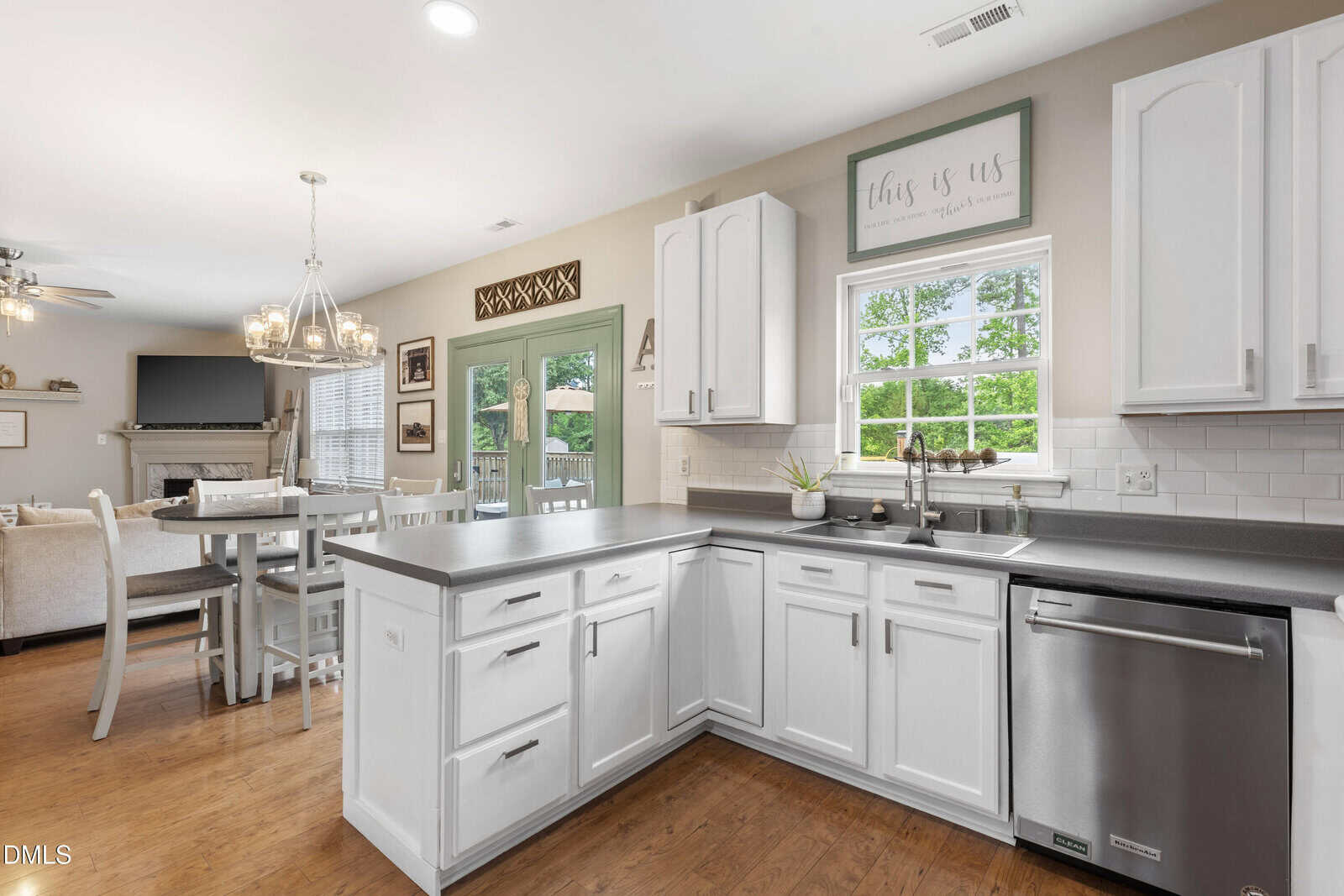 Bright kitchen with white cabinets, stainless steel appliances, gray countertops, and a window above the sink. Dining area with a round table and chairs is visible, along with green double doors leading outside.