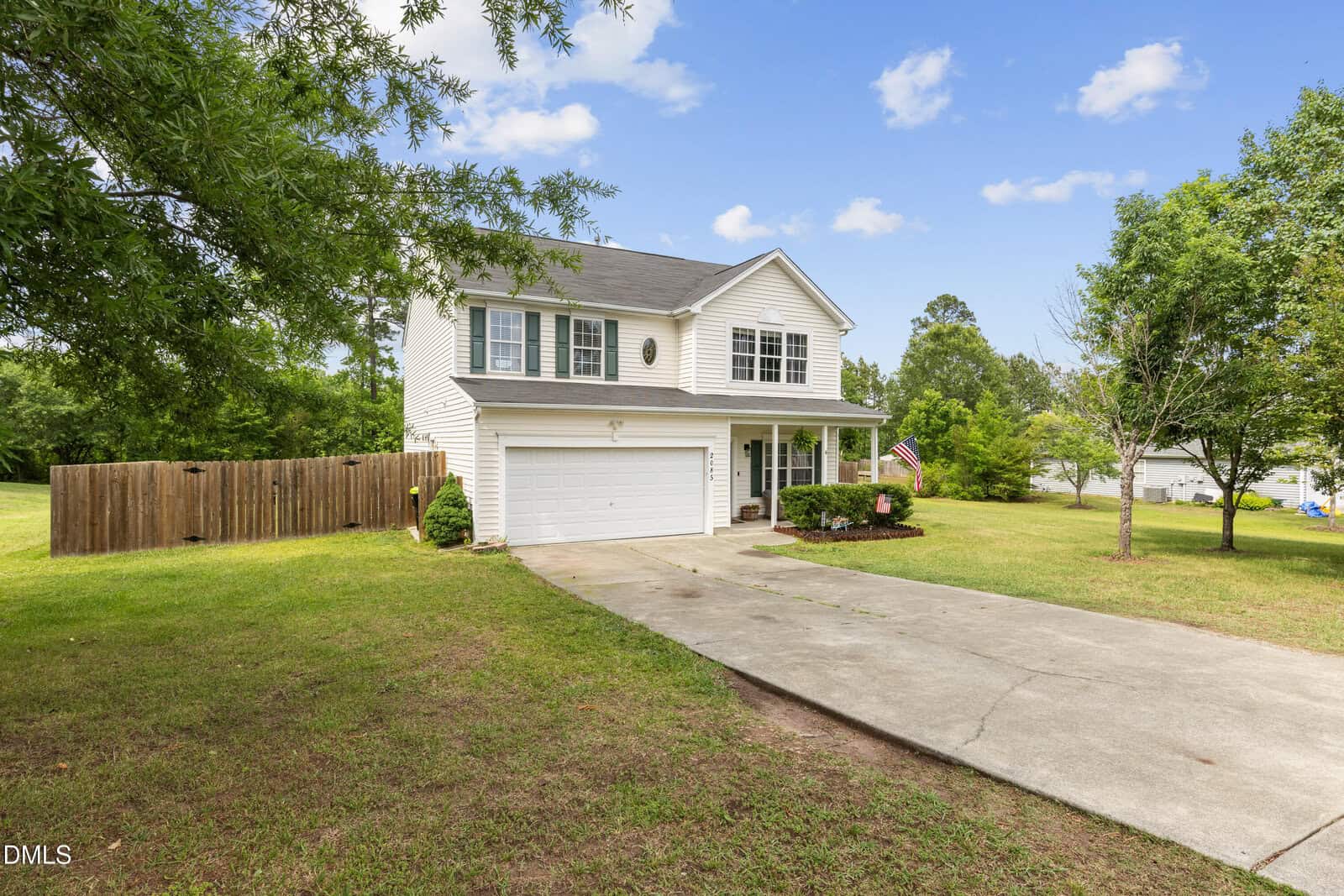 Two-story white house with black shutters and a double garage, set on a spacious grassy lot with trees, a wooden fence on the left, and a concrete driveway leading to the garage under a blue sky with scattered clouds.