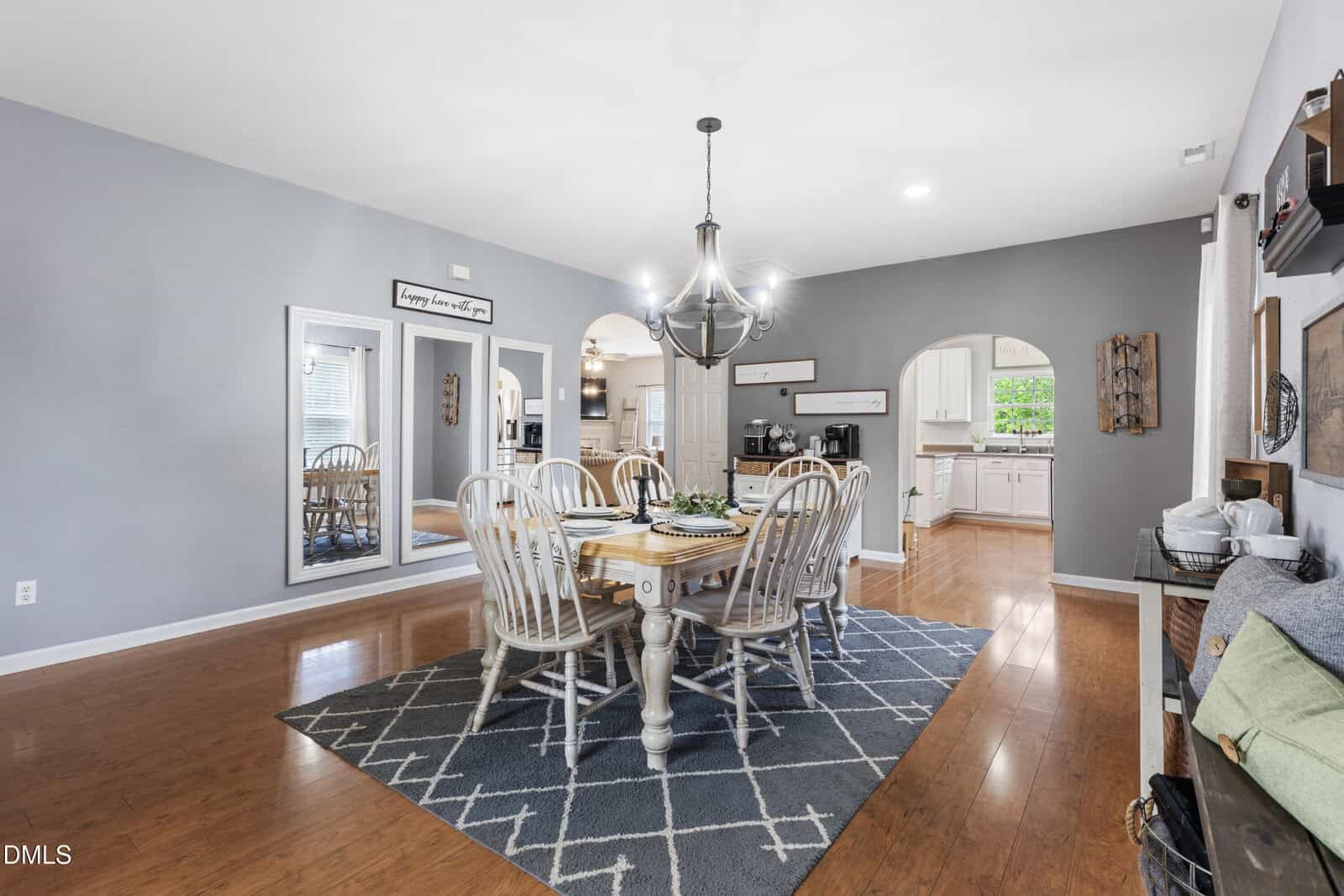 Dining room with six chairs around a wooden table, chandelier above, gray rug, hardwood floors, gray walls, and open archways leading to a kitchen with white cabinets. Large mirrors and wall décor add to the space.