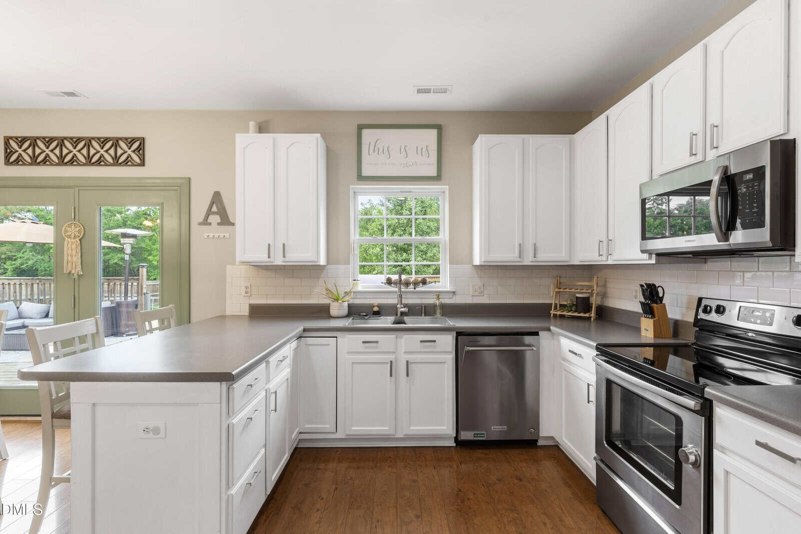 Bright kitchen with white cabinets, gray countertops, stainless steel appliances, potted plants, and a window over the sink. Patio doors on the left open to a deck with outdoor furniture visible outside.