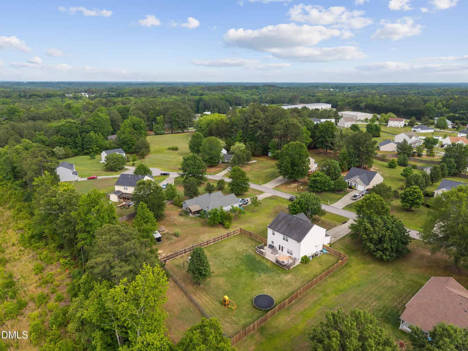 Aerial view of a suburban neighborhood with houses, green lawns, trees, and a fenced backyard featuring a playset and trampoline under a partly cloudy sky.