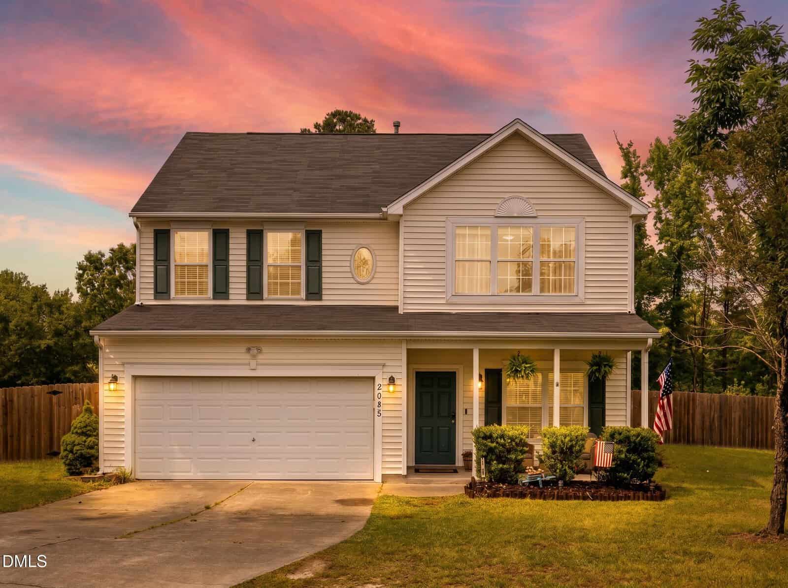 A two-story suburban house at 2085 Partridge Court, Creedmoor, with white siding, a dark roof, and a two-car garage at sunset. The front yard offers bushes, an American flag, and a tree, plus a wooden fence and trees in the background.
