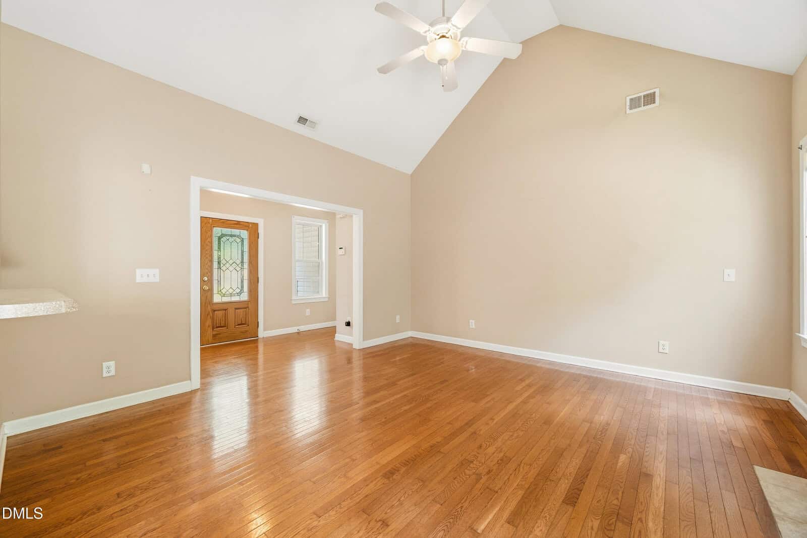 Spacious, empty living room with vaulted ceiling, light beige walls, hardwood floors, ceiling fan with light, and a wooden front door with a decorative glass panel. Large window allows natural light.