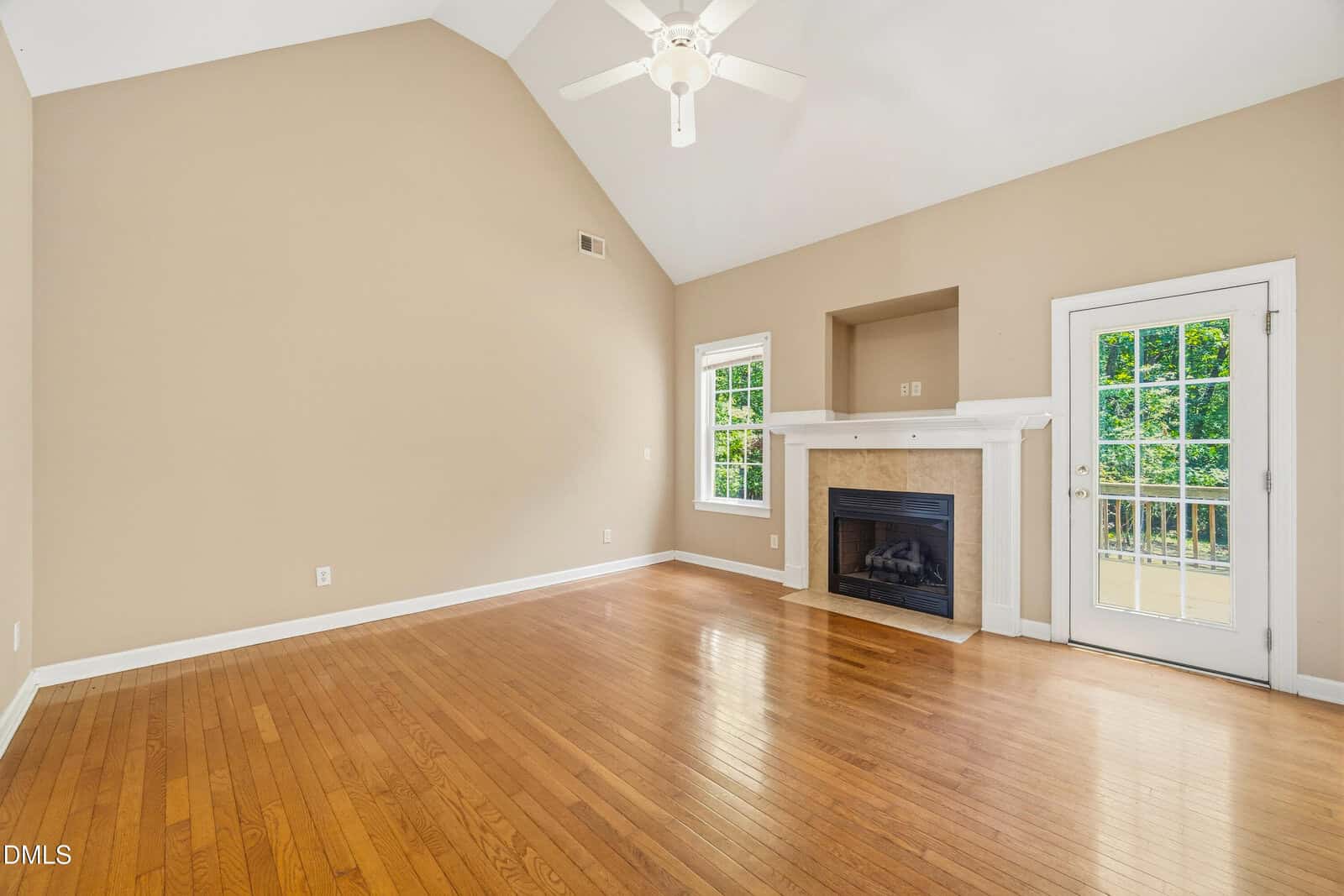 Bright, empty living room with vaulted ceiling, hardwood floors, beige walls, a white ceiling fan, fireplace, single window, and a glass door opening to a balcony with greenery outside.