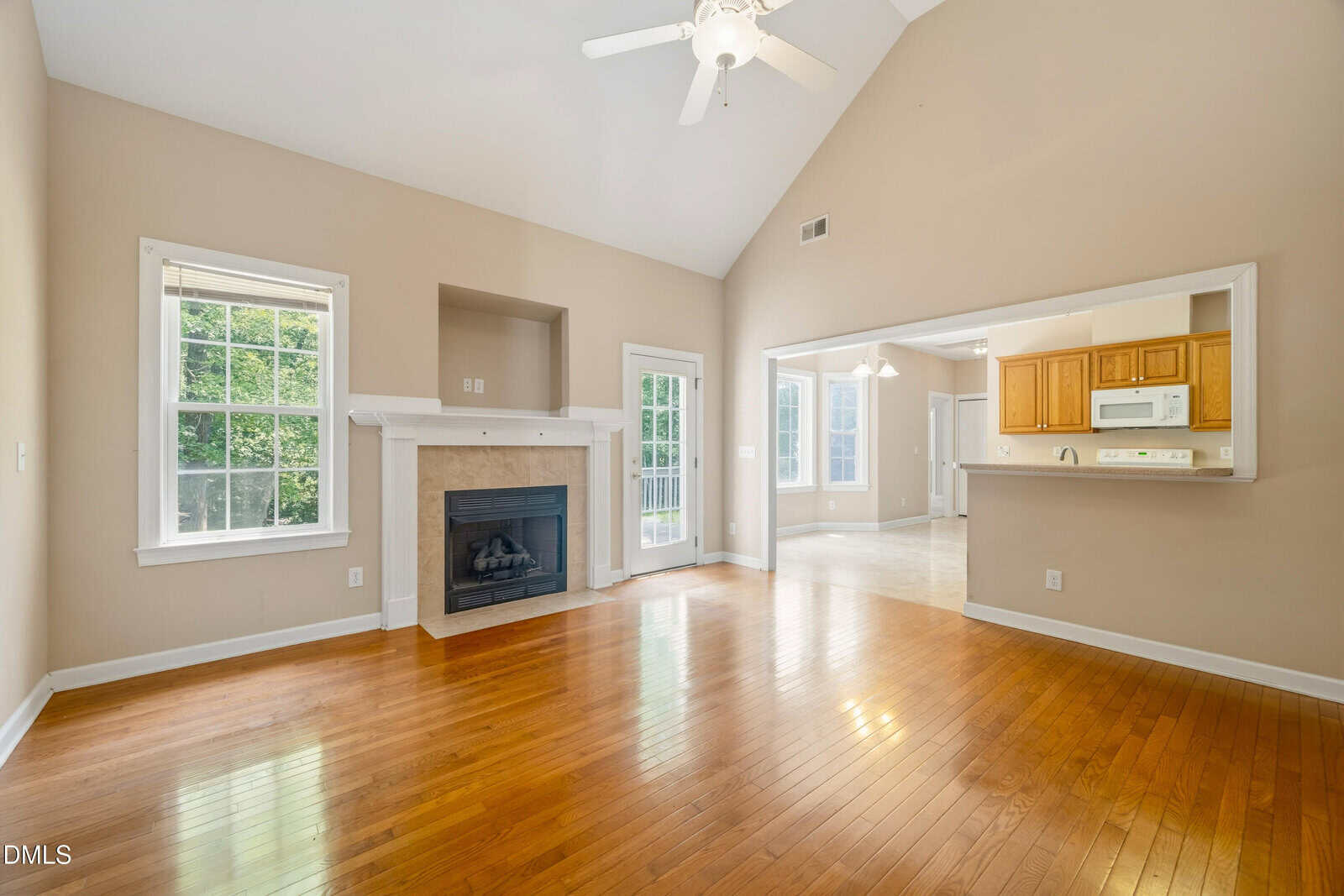 Bright living room with hardwood floors, a ceiling fan, fireplace, and large windows. An open doorway leads to a kitchen with light wood cabinets and white appliances, creating an airy, connected space.