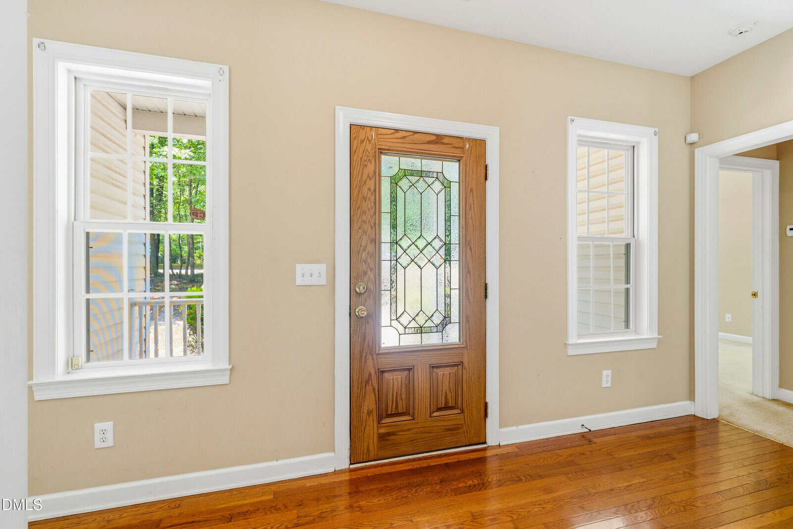 A wooden front door with decorative glass panels is flanked by two white-trimmed windows. The room has beige walls, hardwood floors, and natural light coming through the windows. A hallway is visible on the right.