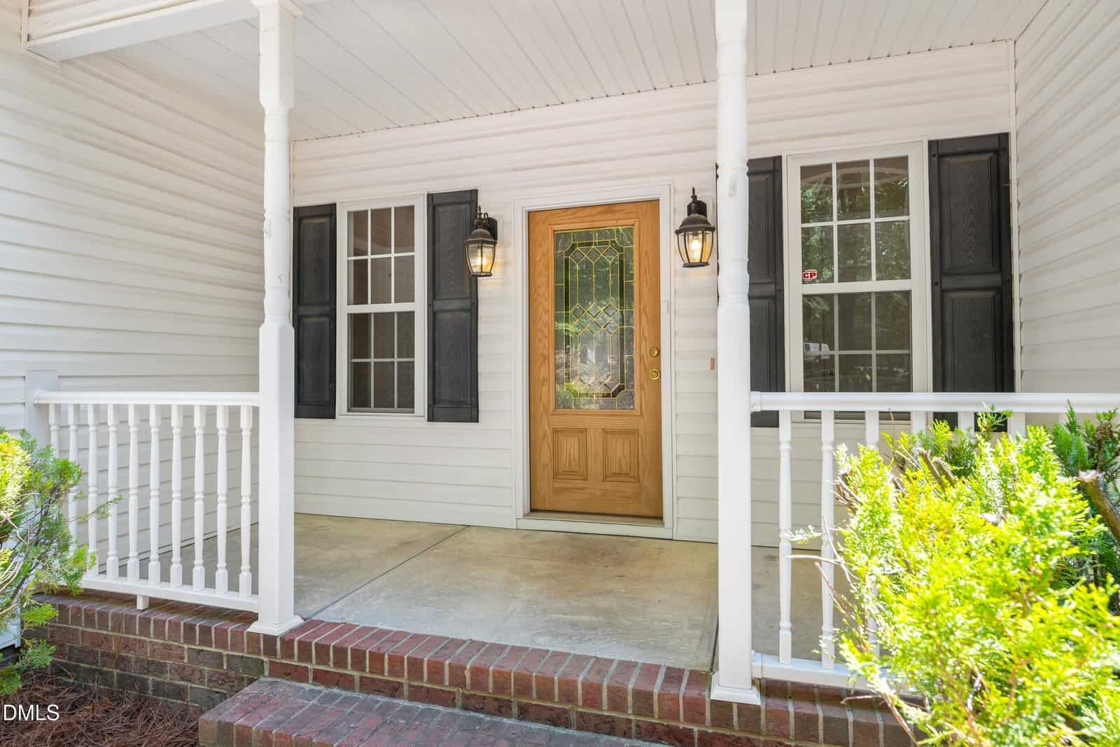 A covered front porch with white railings, a wooden door with decorative glass, two black lantern-style lights, and windows with black shutters on a white house.