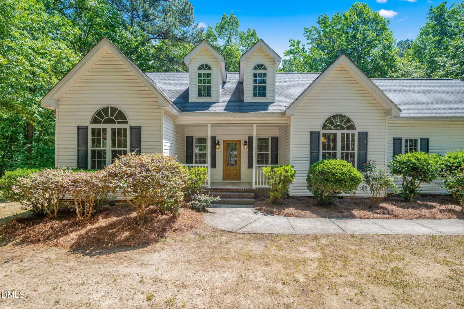 A white single-story house with two dormer windows, black shutters, a covered front porch with wooden door, and neatly trimmed bushes along a curved walkway, surrounded by trees.