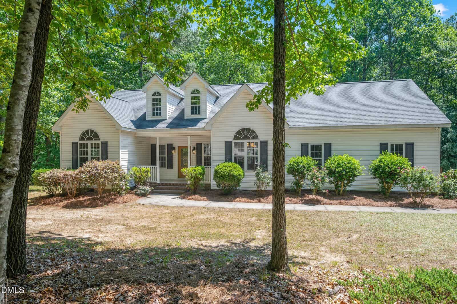 A white single-story house with dormer windows at 675 Timberlands Drive, Louisburg, features a covered front porch and shrubs, surrounded by trees and greenery on a sunny day.