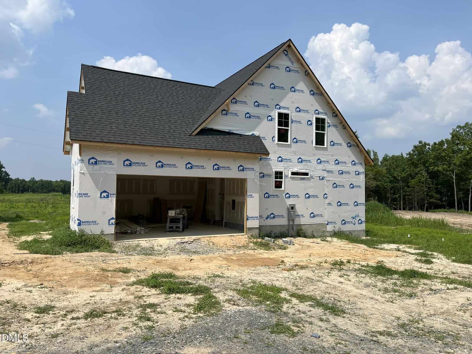 A partially constructed house wrapped in white weatherproofing material sits on a dirt lot. The garage door is open, revealing construction materials inside. The yard is unfinished, and trees are visible in the background.