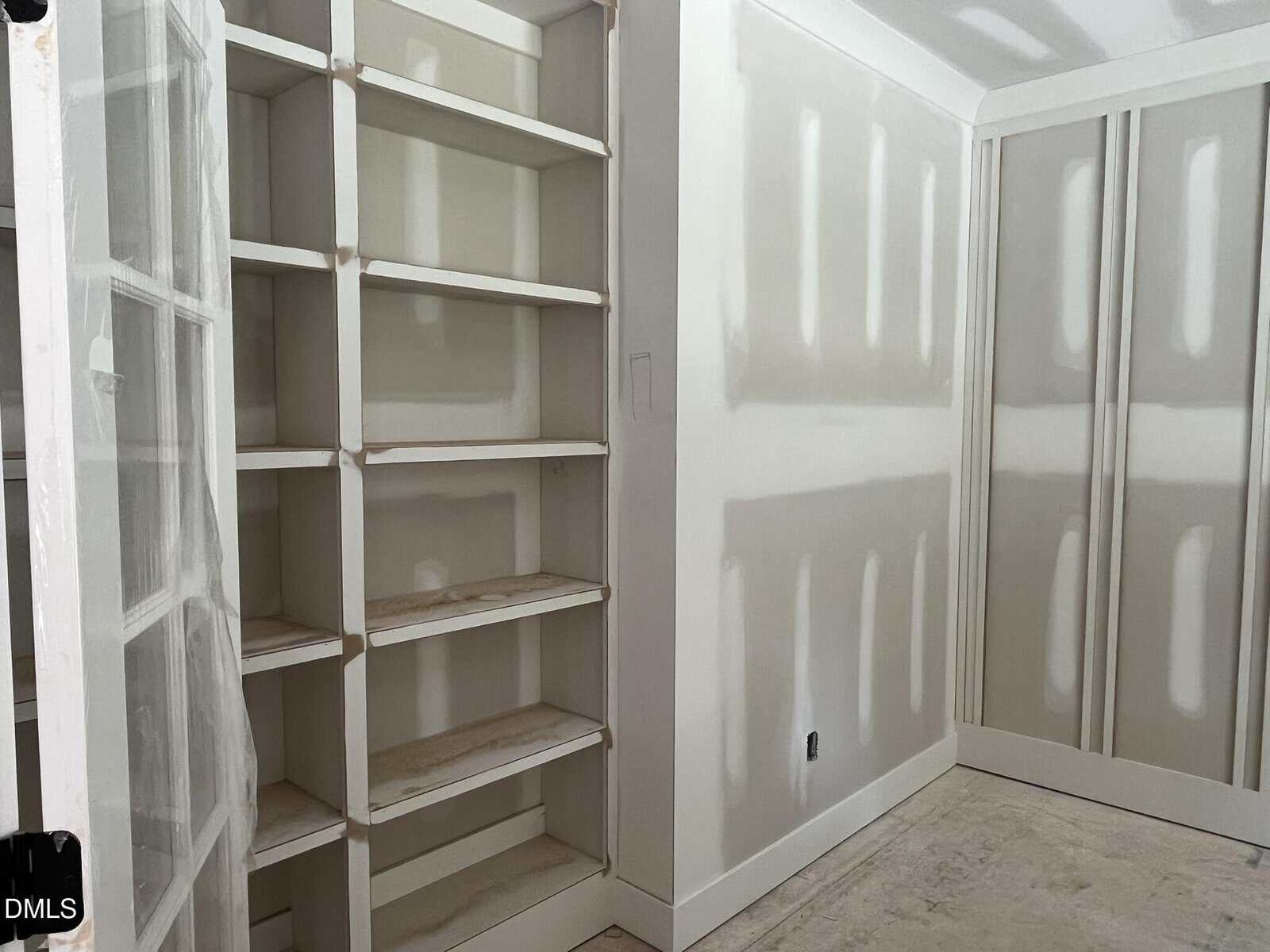 A room under construction with unfinished drywall, built-in white shelves, and closed double doors. The floor and shelves have dust and some construction debris.