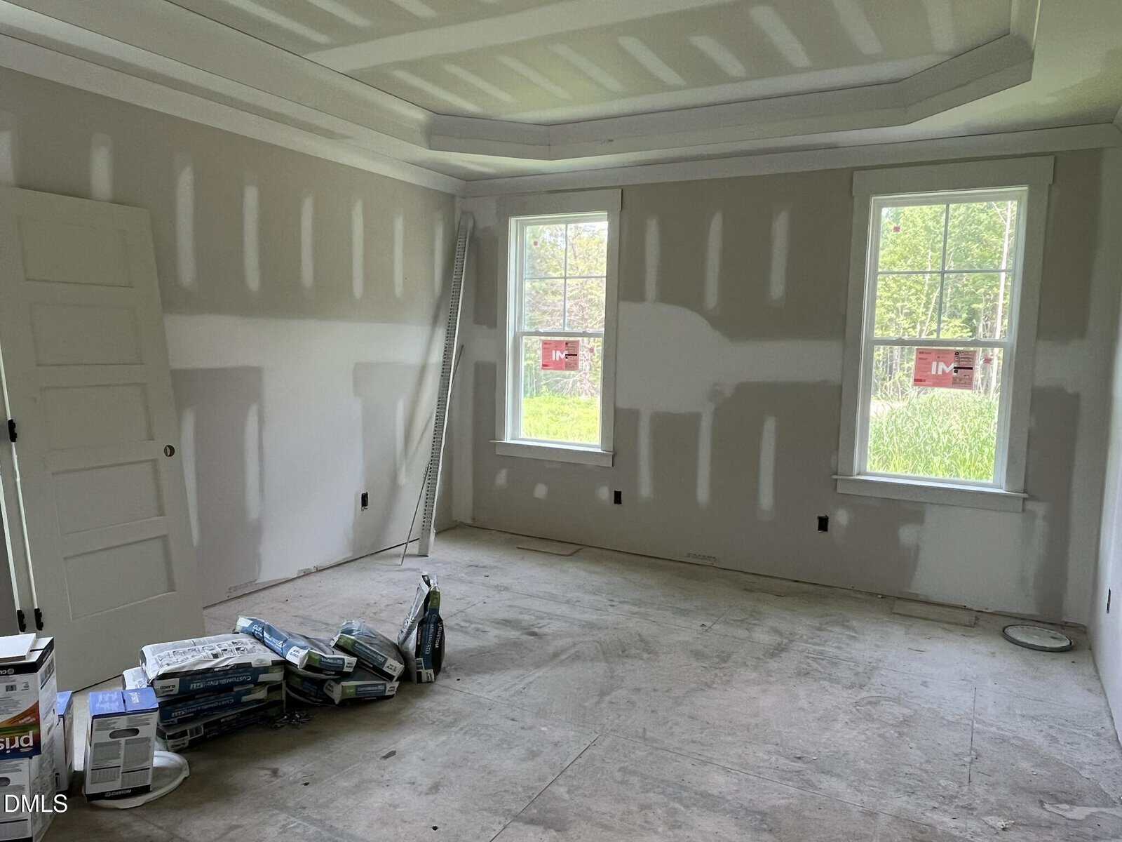 An unfinished room with drywall installed but not painted, construction materials and tools on the floor, an open door, and two large windows letting in natural light.
