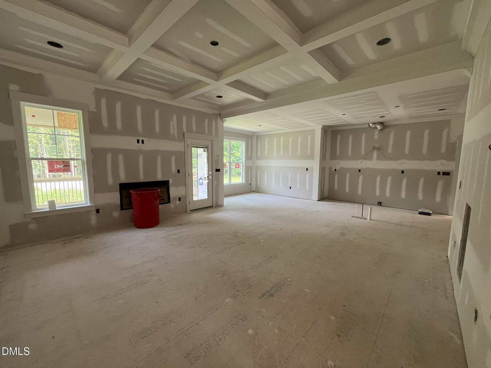 Unfinished room with drywall installed, unpainted walls, coffered ceiling, fireplace, large windows, glass doors, and a red trash bin near the corner. Room is empty and well-lit with natural light.