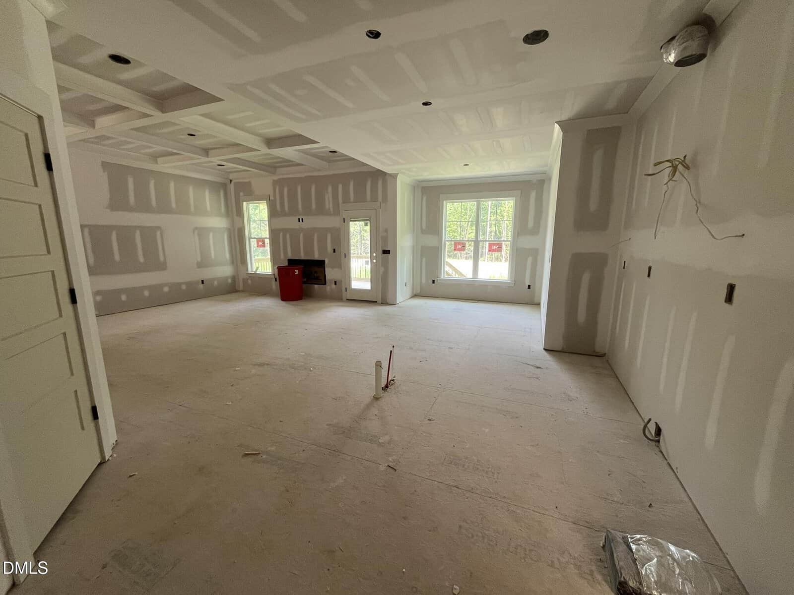 Large, unfinished room with drywall installation in progress, exposed joints, and an unpainted floor. Sunlight streams through large windows and a door at the back. Some construction materials are visible.